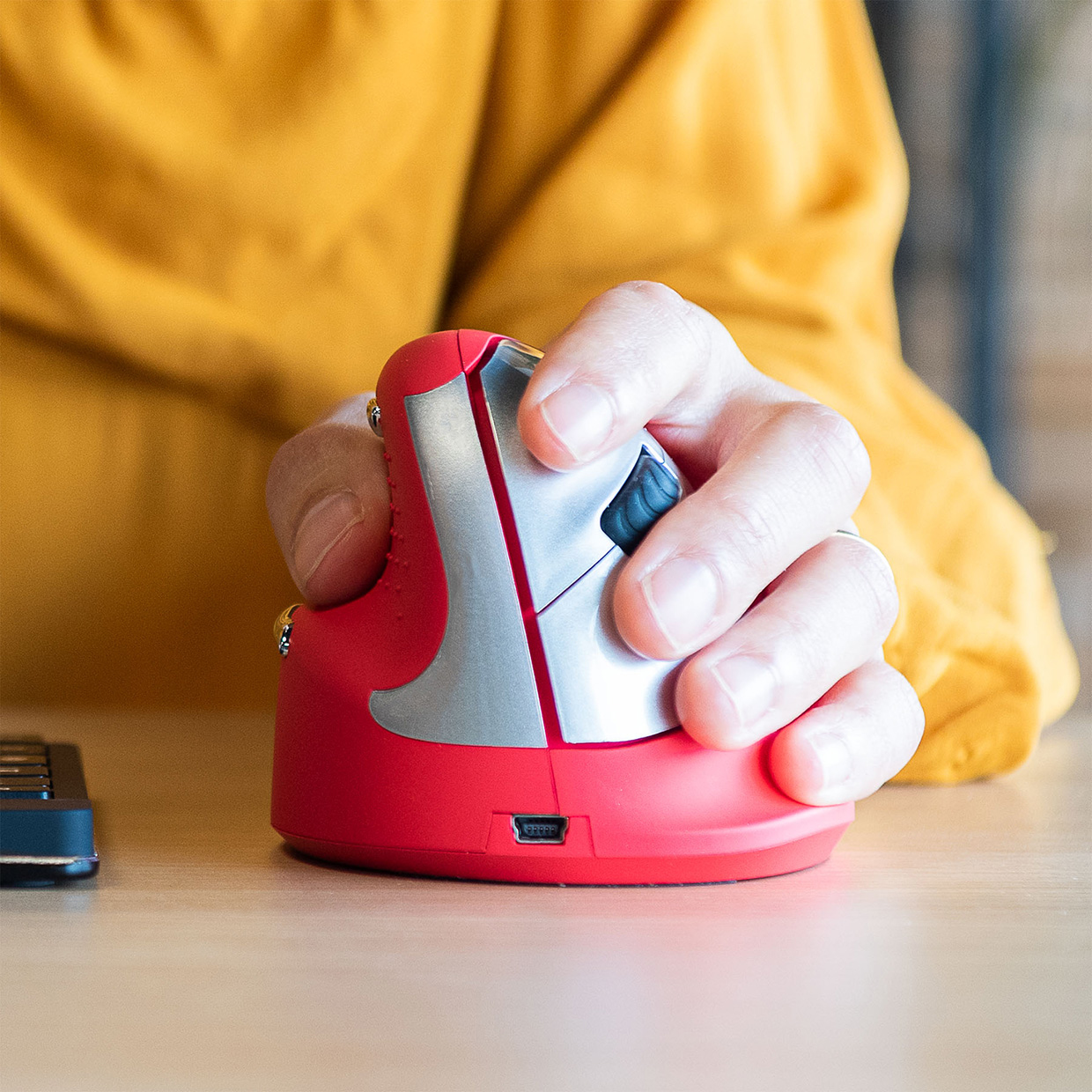 Mano sujetando ratón ergonómico rojo. Se ve camisa amarilla y escritorio.