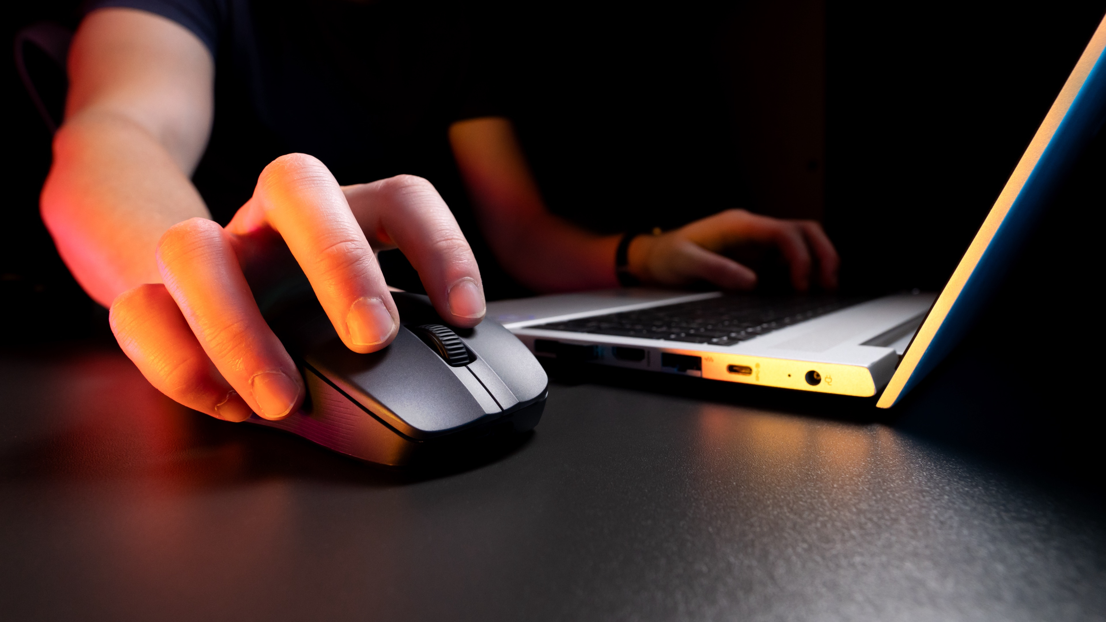 Dłoń używająca czarnej myszy na ciemnej powierzchni, z podświetlonym laptopem w tle.