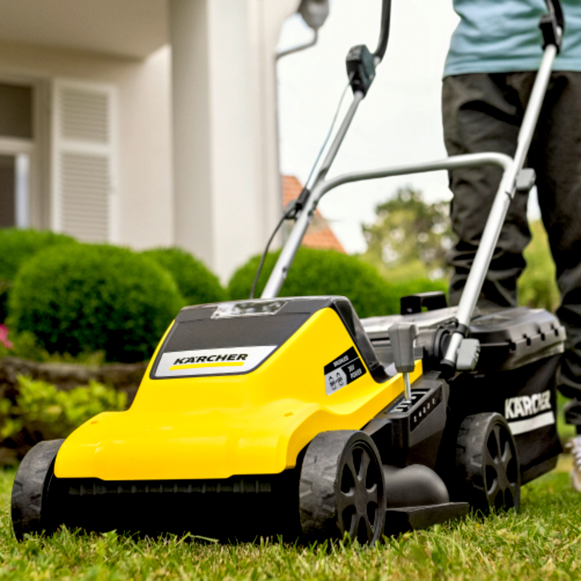 Gelb-schwarzer Karcher-Rasenmäher auf grünem Gras, eine Person mäht den Rasen vor einem Haus.