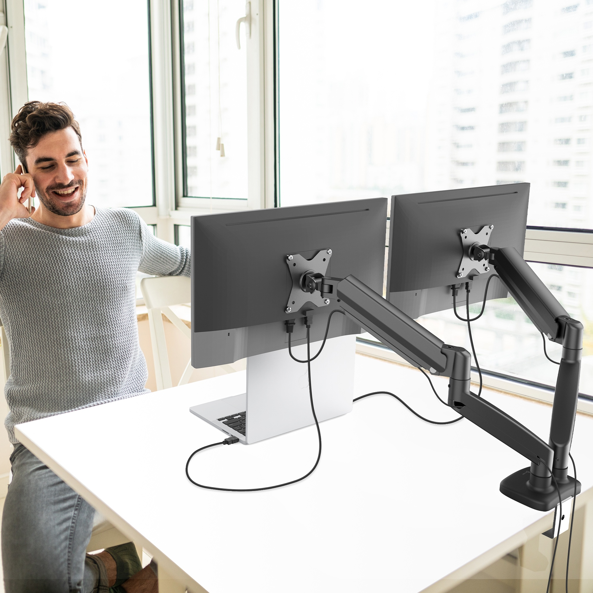 Homme au téléphone près de deux moniteurs sur un bureau blanc. Ordinateur portable et fenêtre en arrière-plan.