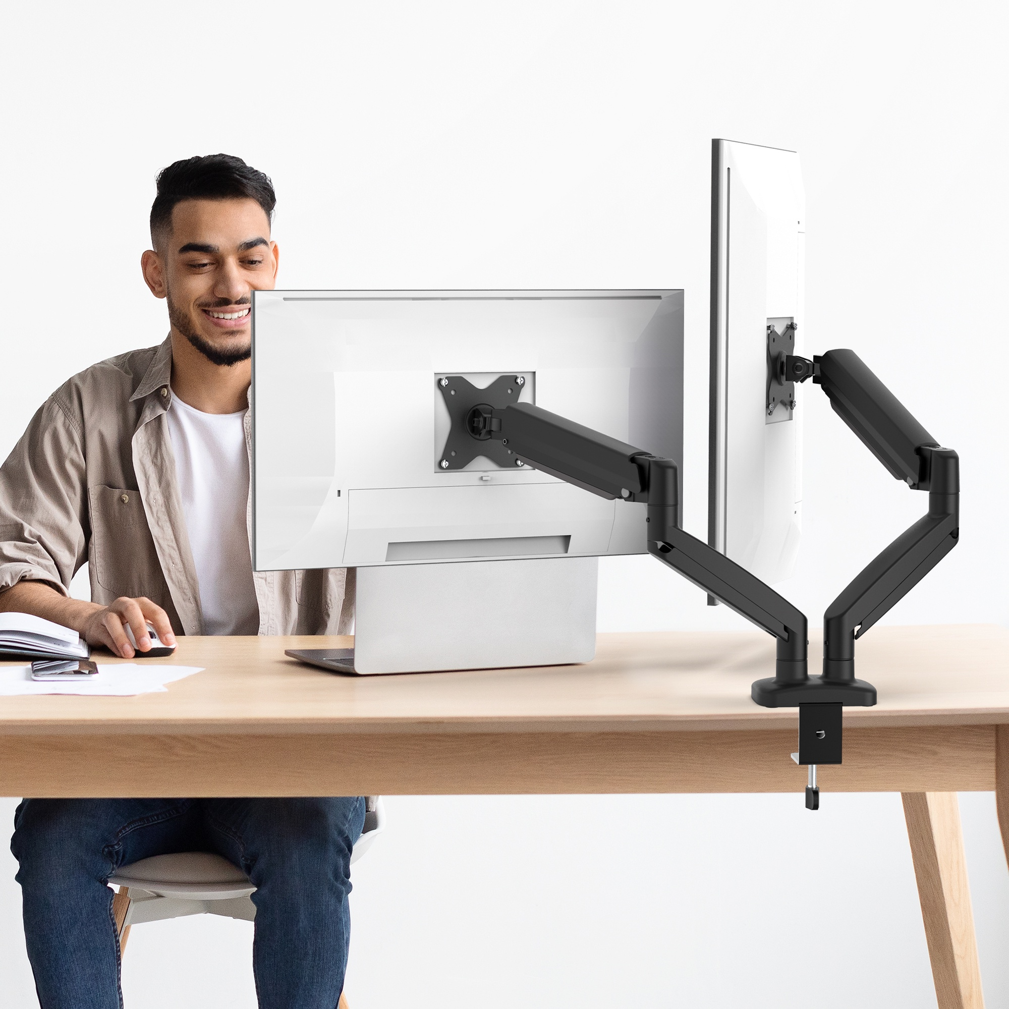 Un homme est assis à un bureau avec deux moniteurs attachés à un bras de moniteur. Le fond est blanc.