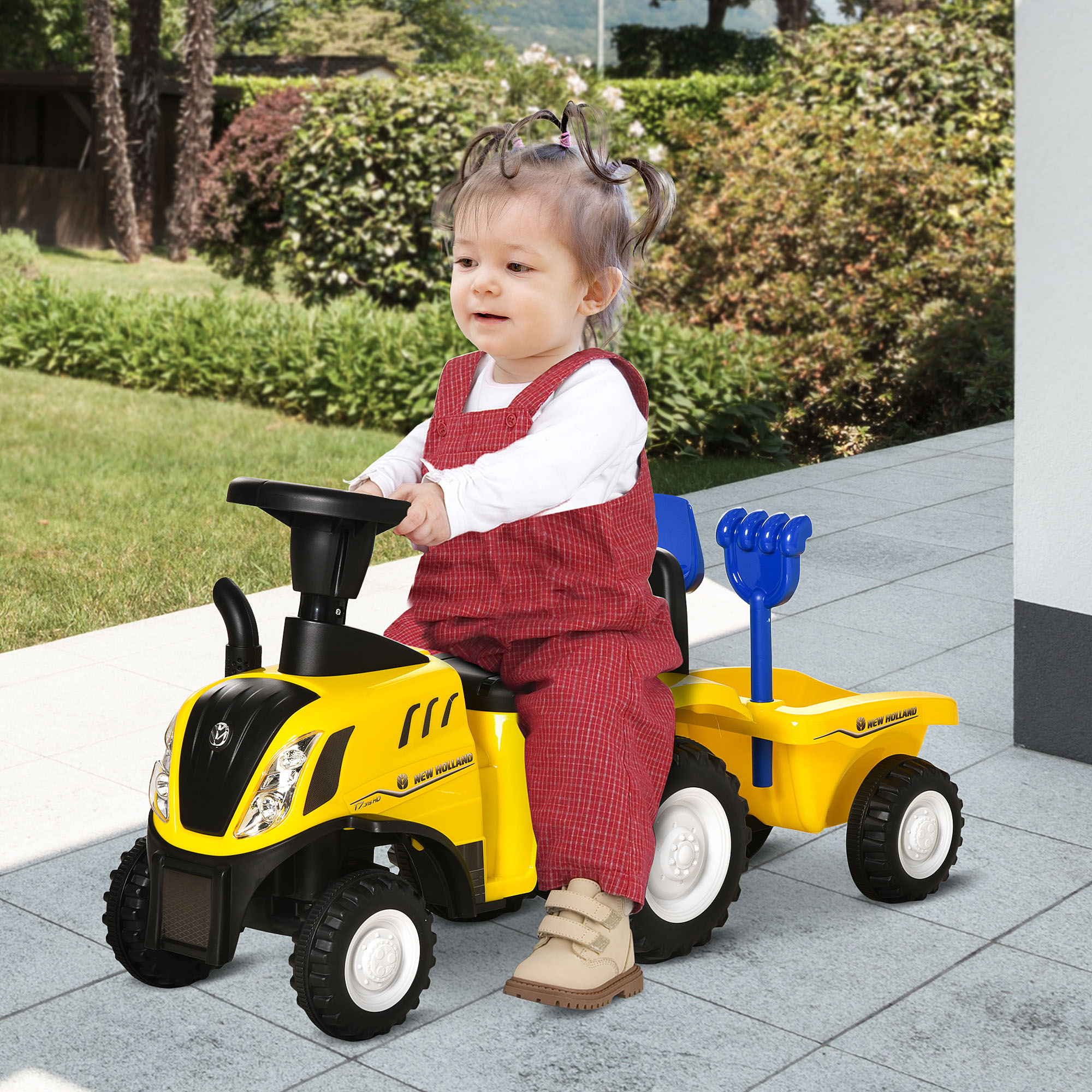 Un bambino piccolo con una tuta rossa guida un trattore giocattolo giallo su un patio. Cespugli verdi sullo sfondo.