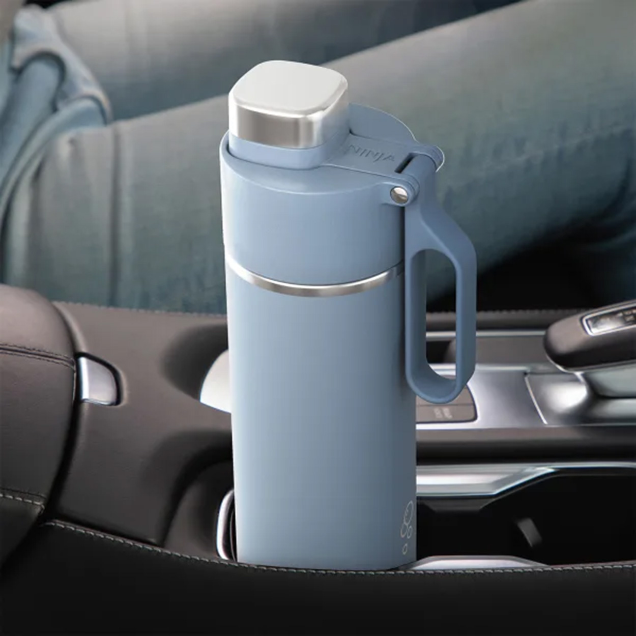 Eine blaue Wasserflasche mit Griff befindet sich in einem Auto-Getränkehalter, in der Nähe der Beine einer Person.