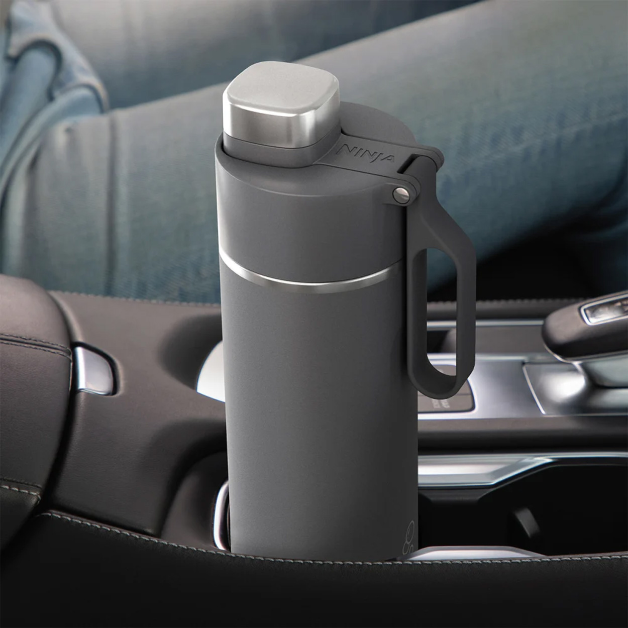 Graue Ninja-Becherflasche mit Griff, in einer Autokonsole platziert, Jeans einer Person im Hintergrund.