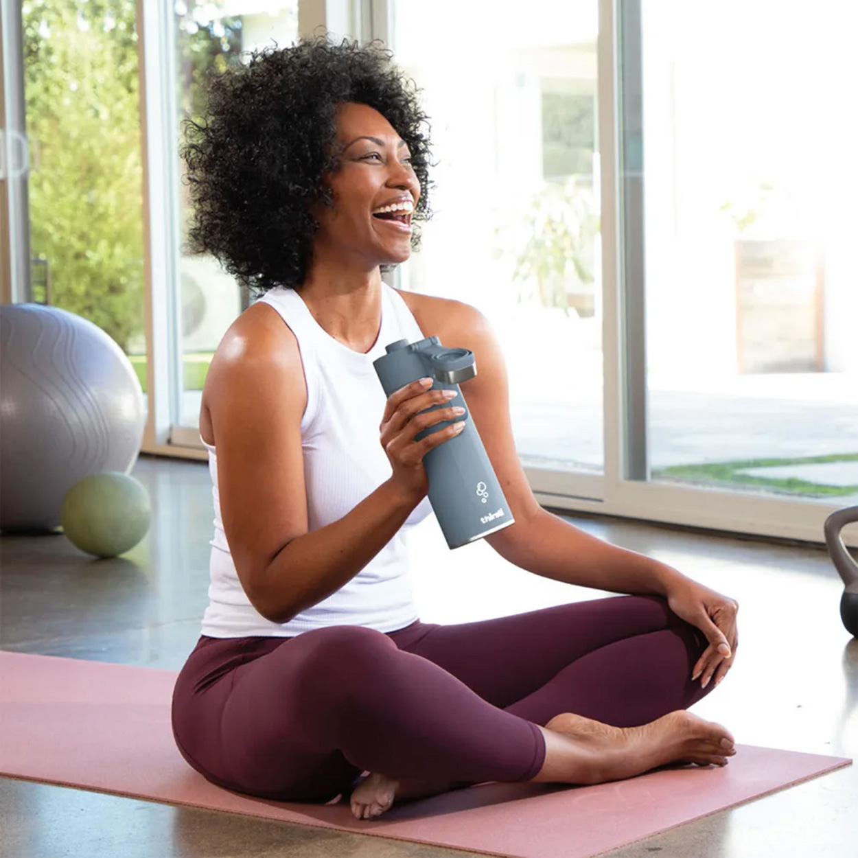 Donna che ride mentre tiene una bottiglia d'acqua grigia, seduta a gambe incrociate su un tappetino da yoga, stanza con porte a vetri.