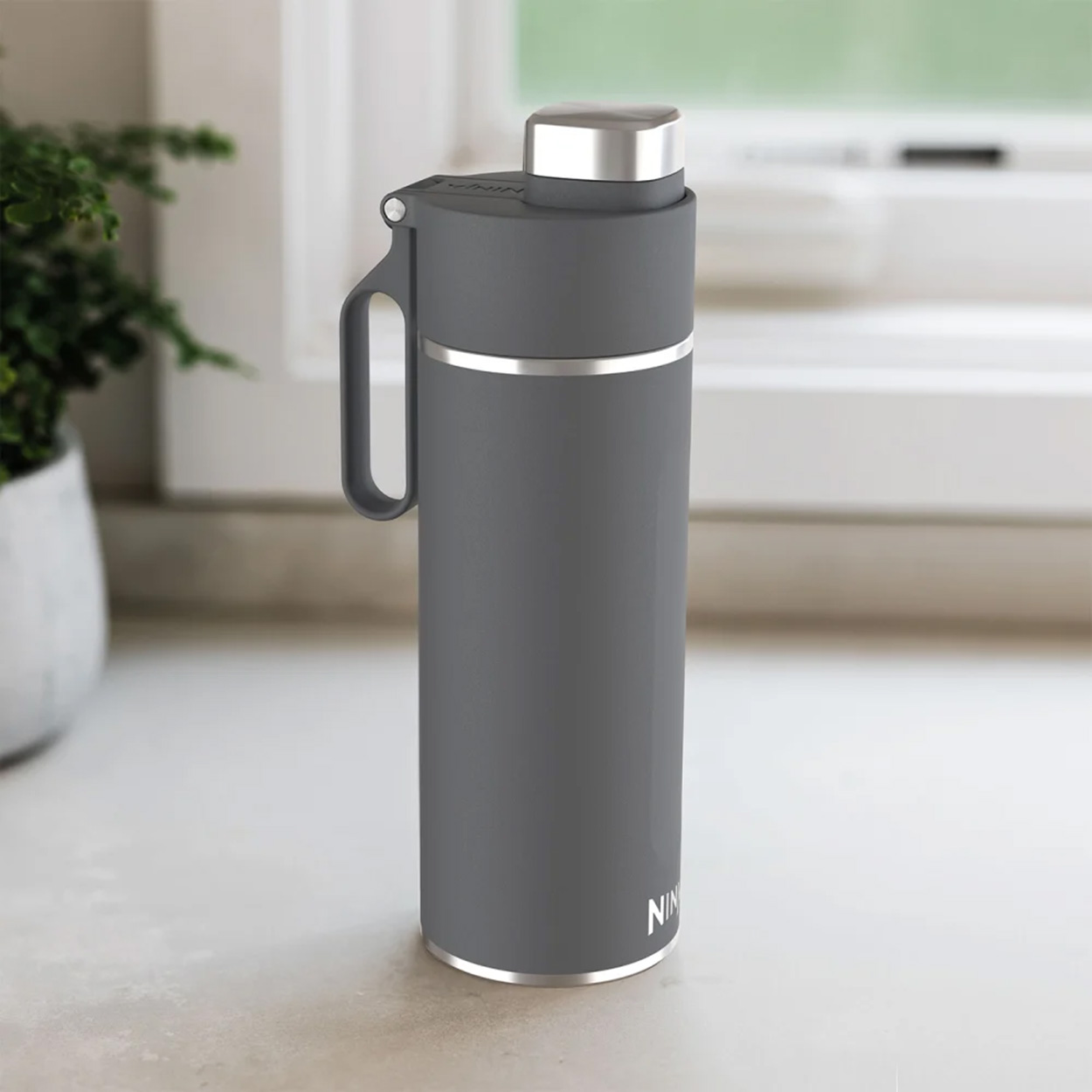 Graue isolierte Wasserflasche mit Griff und silbernem Deckel. Hintergrund: Fenster und Pflanze.