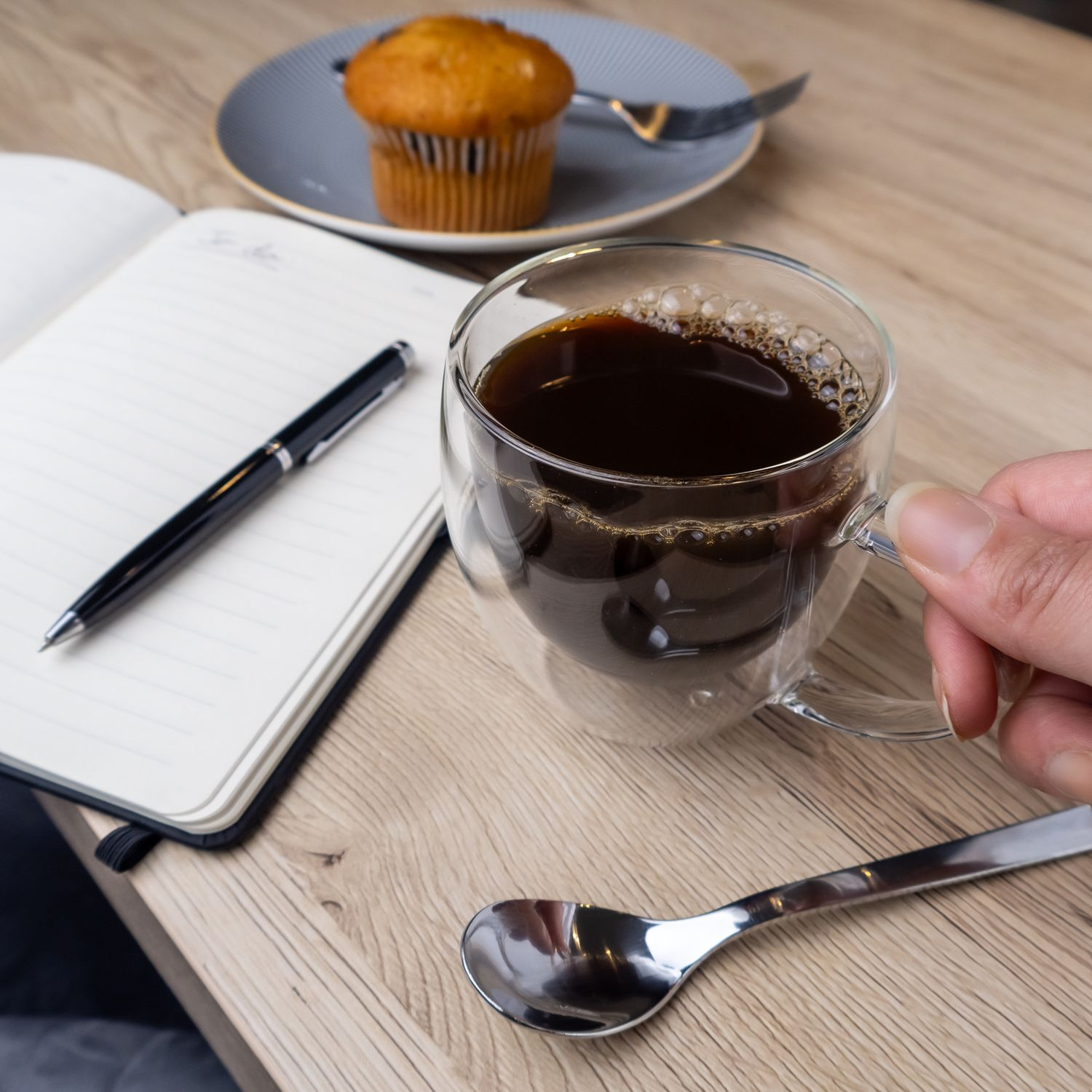 Tasse Kaffee, Notizbuch, Stift und Löffel auf einem Holztisch mit einem Muffin auf einem Teller.