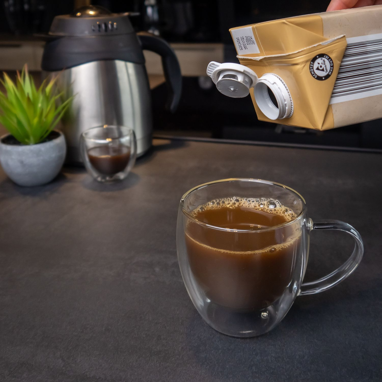 Kaffee wird in einen Glasbecher gegossen. Ein Wasserkocher und eine Pflanze sind im Hintergrund.