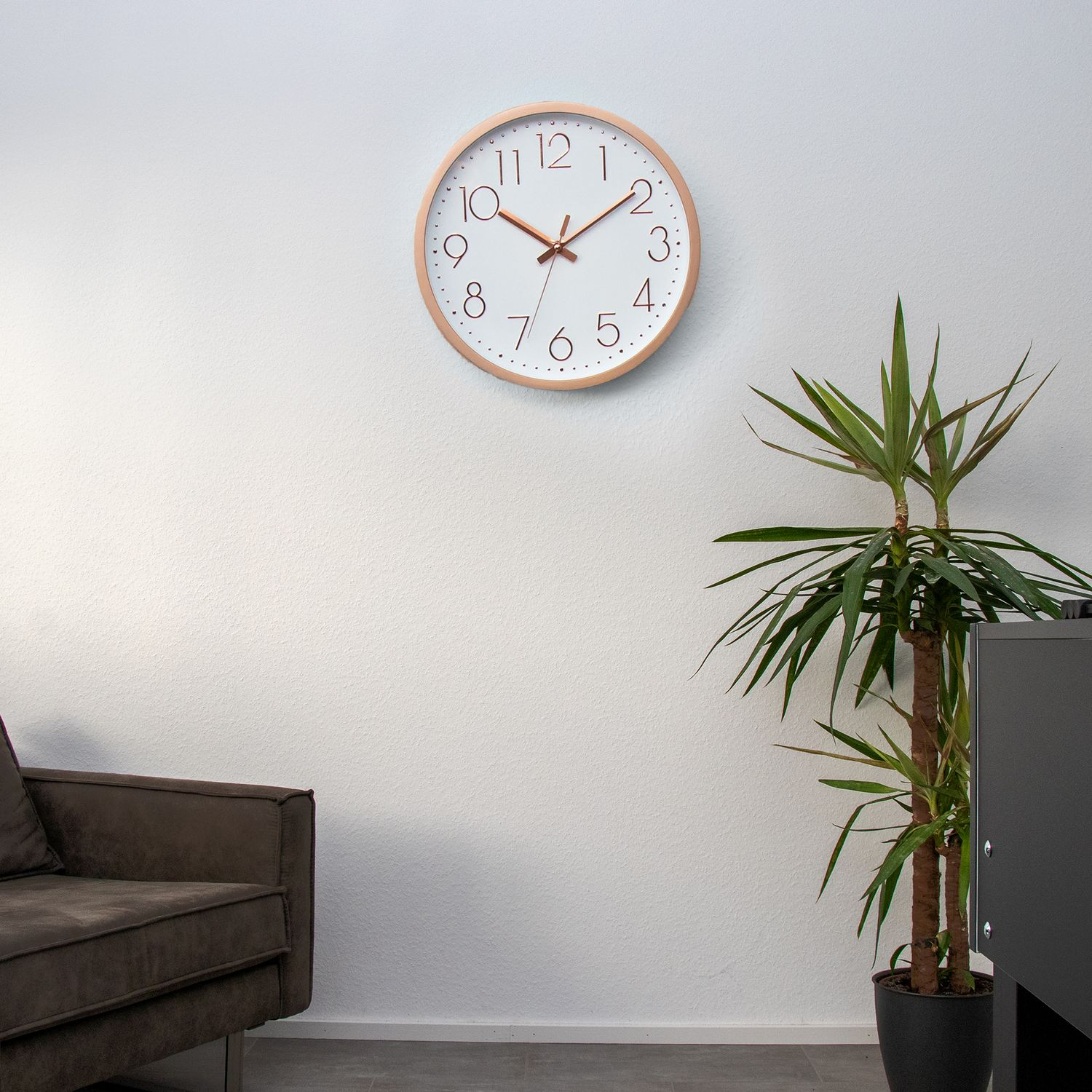 Eine Wanduhr mit hellem Holzrahmen an einer weißen Wand. Daneben steht eine Pflanze.