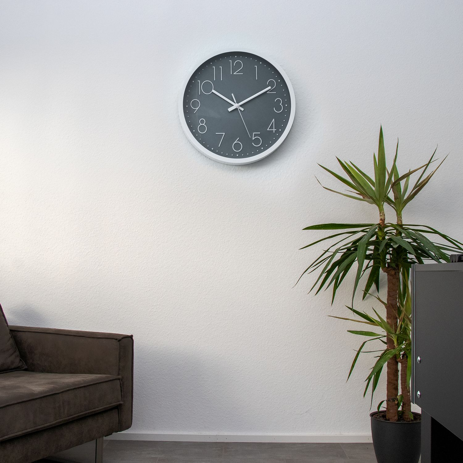 Eine weiße Wanduhr mit schwarzen Zahlen hängt über einem braunen Sofa und einer Pflanze.