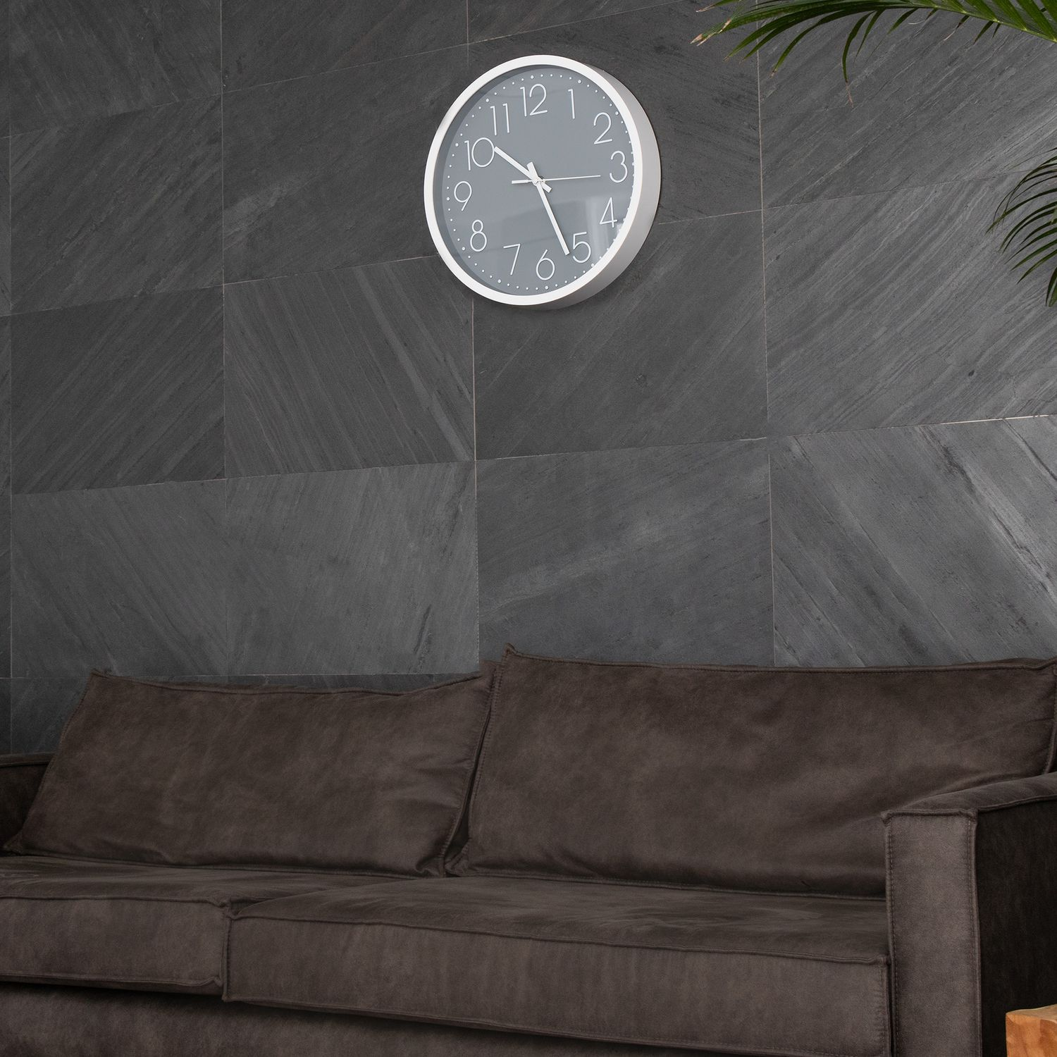 Weiße Uhr an grauer Wand über braunem Sofa. Eine Pflanze in der rechten Ecke.