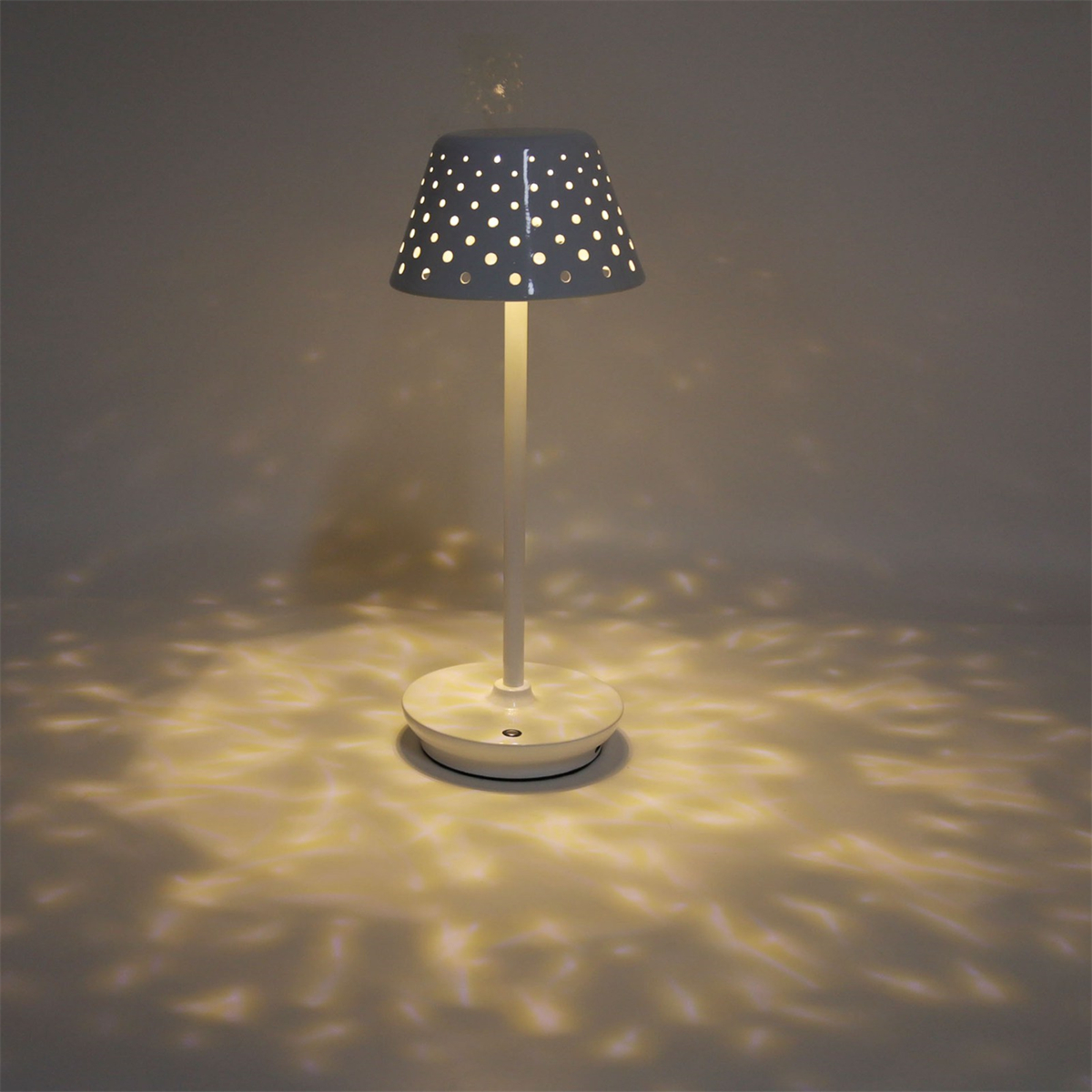 Eine weiße Tischlampe mit perforiertem Schirm. Die Lampe wirft Licht auf den Boden. Der Hintergrund ist grau.