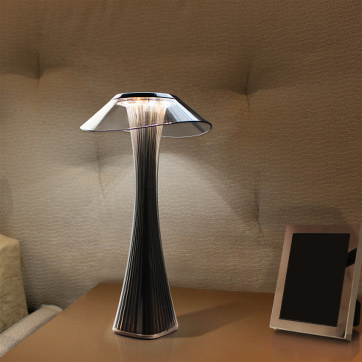 Moderne Lampe mit schwarzem Sockel und transparentem Schirm, auf einem Holztisch. Rechts ein Rahmen.