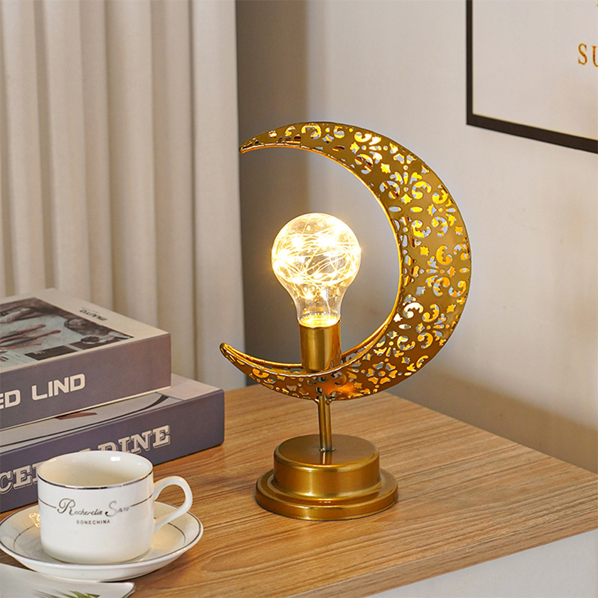Eine goldene mondförmige Lampe mit Glühbirne, die auf einem Holztisch steht, neben einer Kaffeetasse und Büchern.
