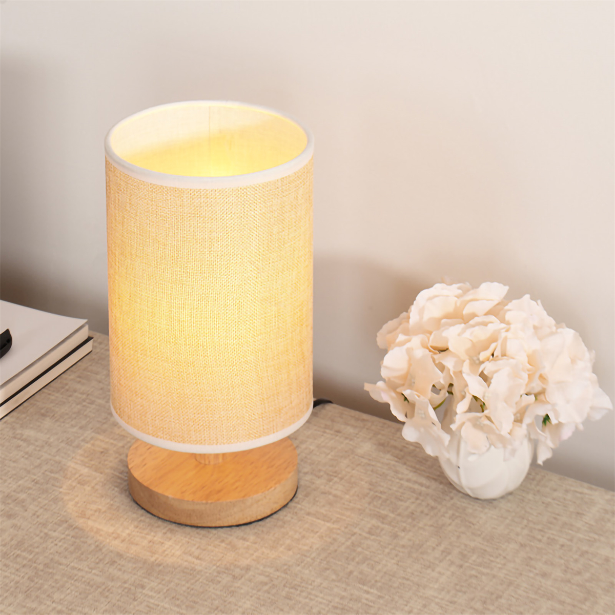 Beige Stofflampe mit Holzsockel und weißen Blumen auf einem Tisch.