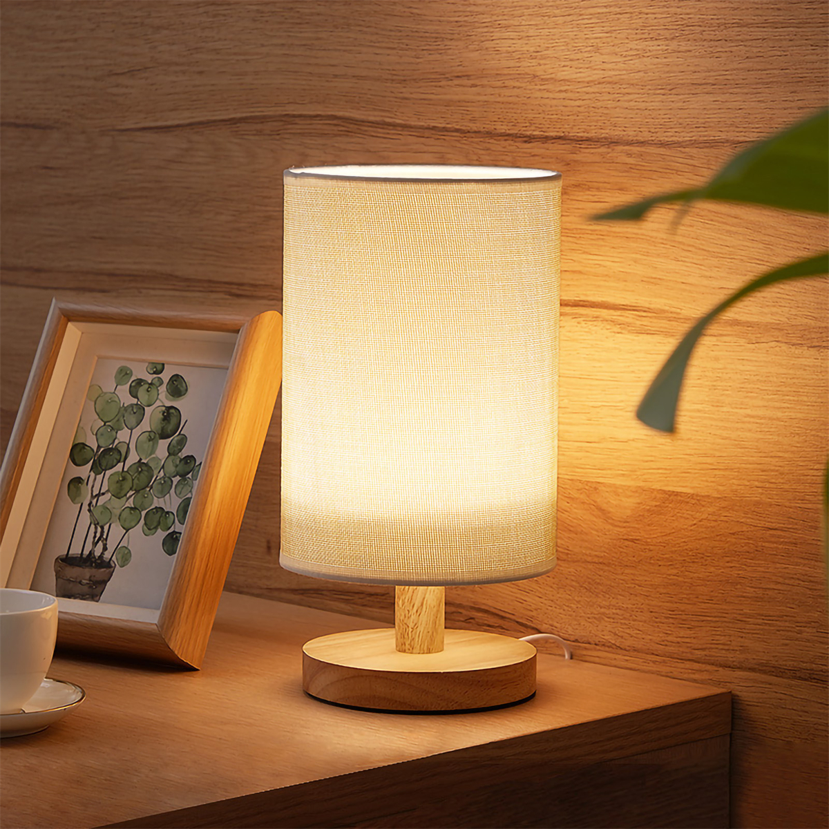 Beleuchtete Tischlampe mit beigem Schirm auf einem Holztisch. Ein gerahmtes Bild und eine Tasse.