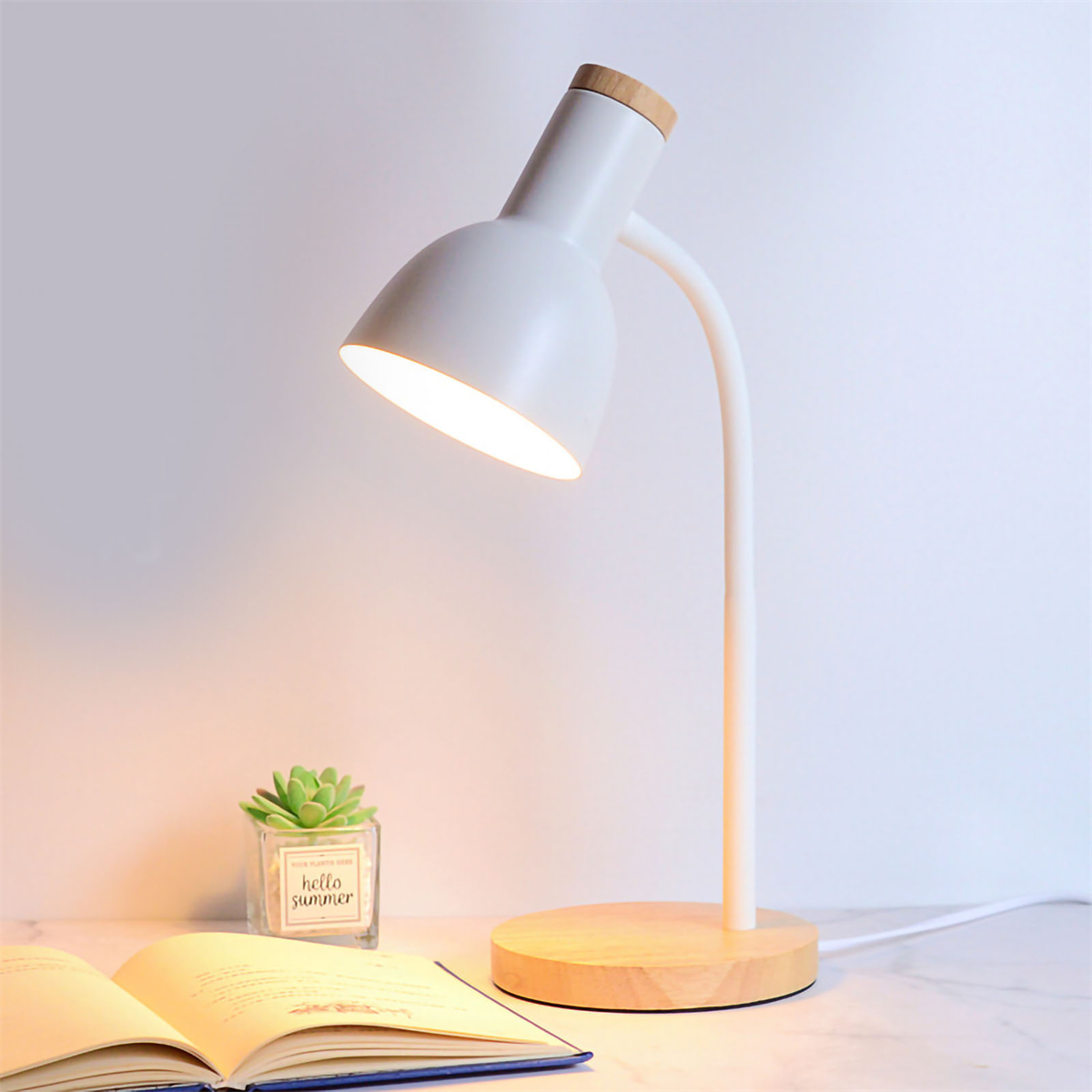 Eine weiße Schreibtischlampe mit Holzsockel, die ein Buch und eine kleine Pflanze in einem durchsichtigen Topf beleuchtet.