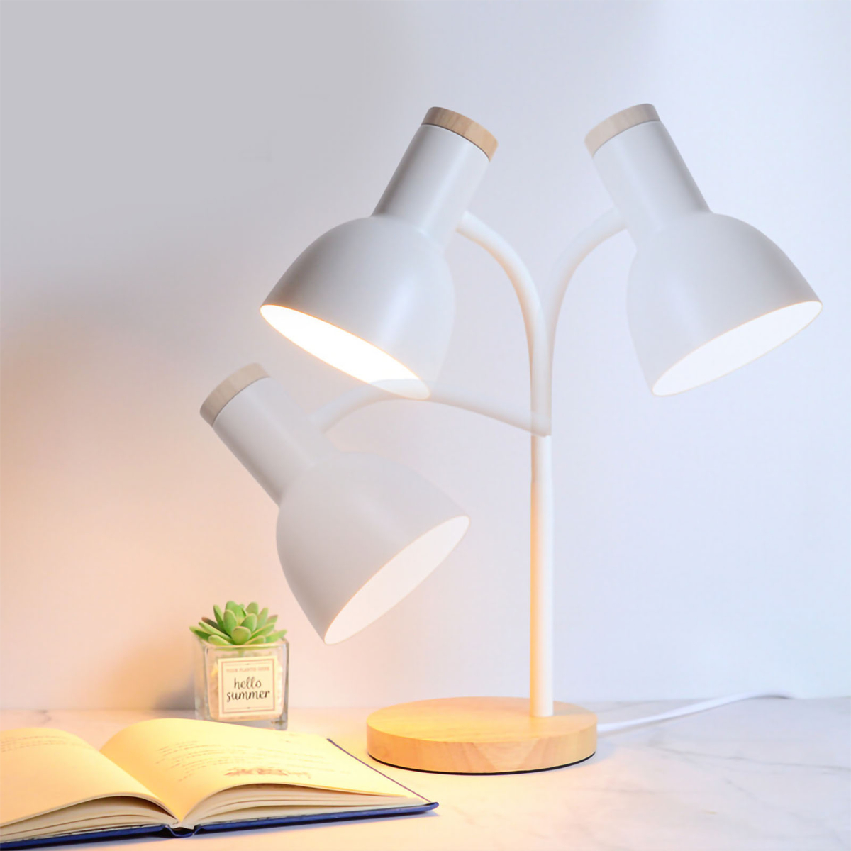 Eine weiße Lampe mit drei verstellbaren Köpfen auf einem Holzsockel, neben einem Buch und einer Pflanze.