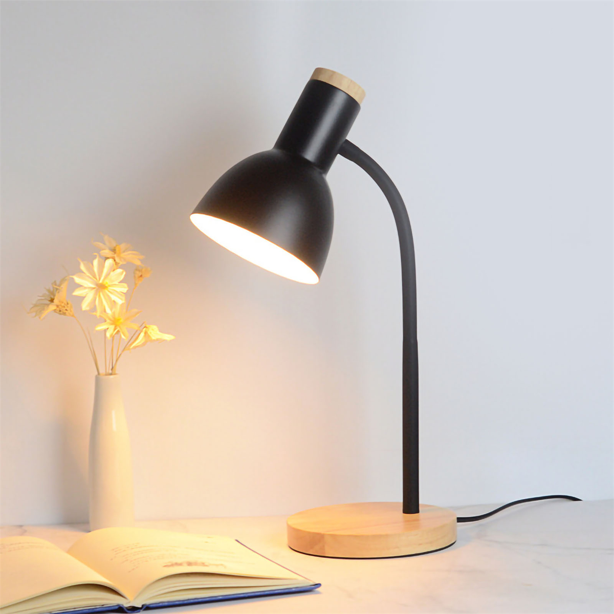 Schwarze Schreibtischlampe mit Holzfuß, Blumenvase und Buch auf einem Tisch.