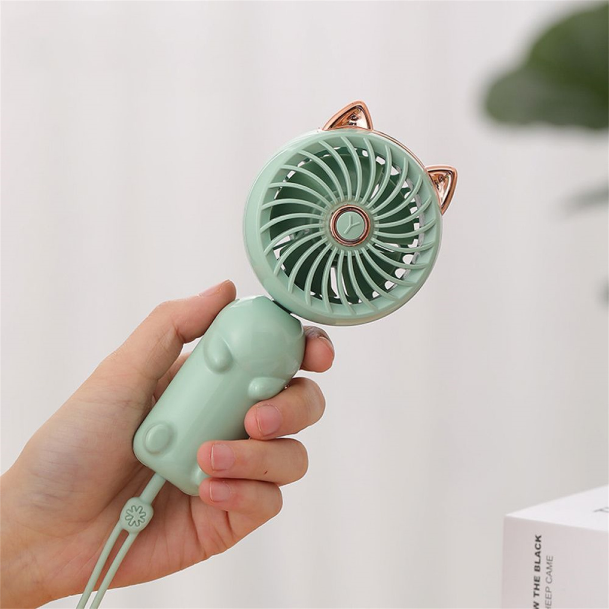 Ein grüner Fächer mit Katzenohren. Hand, die den Ventilator hält. Der Hintergrund ist weiß.