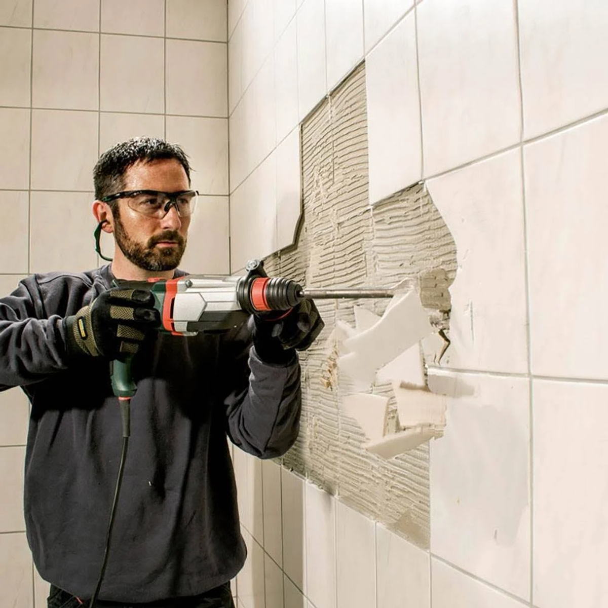 Mężczyzna wierci w ścianie. Okulary ochronne i ochrona słuchu. Biała ściana z płytek.