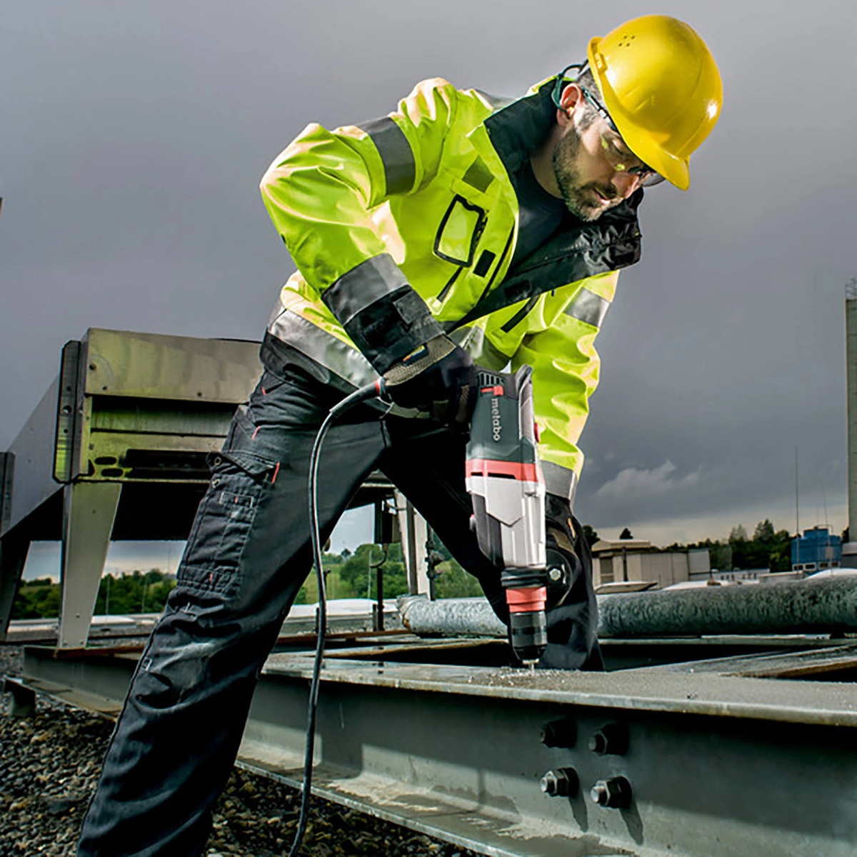 Osoba w odzieży roboczej wierci w metalu. Scena ma pochmurne tło.