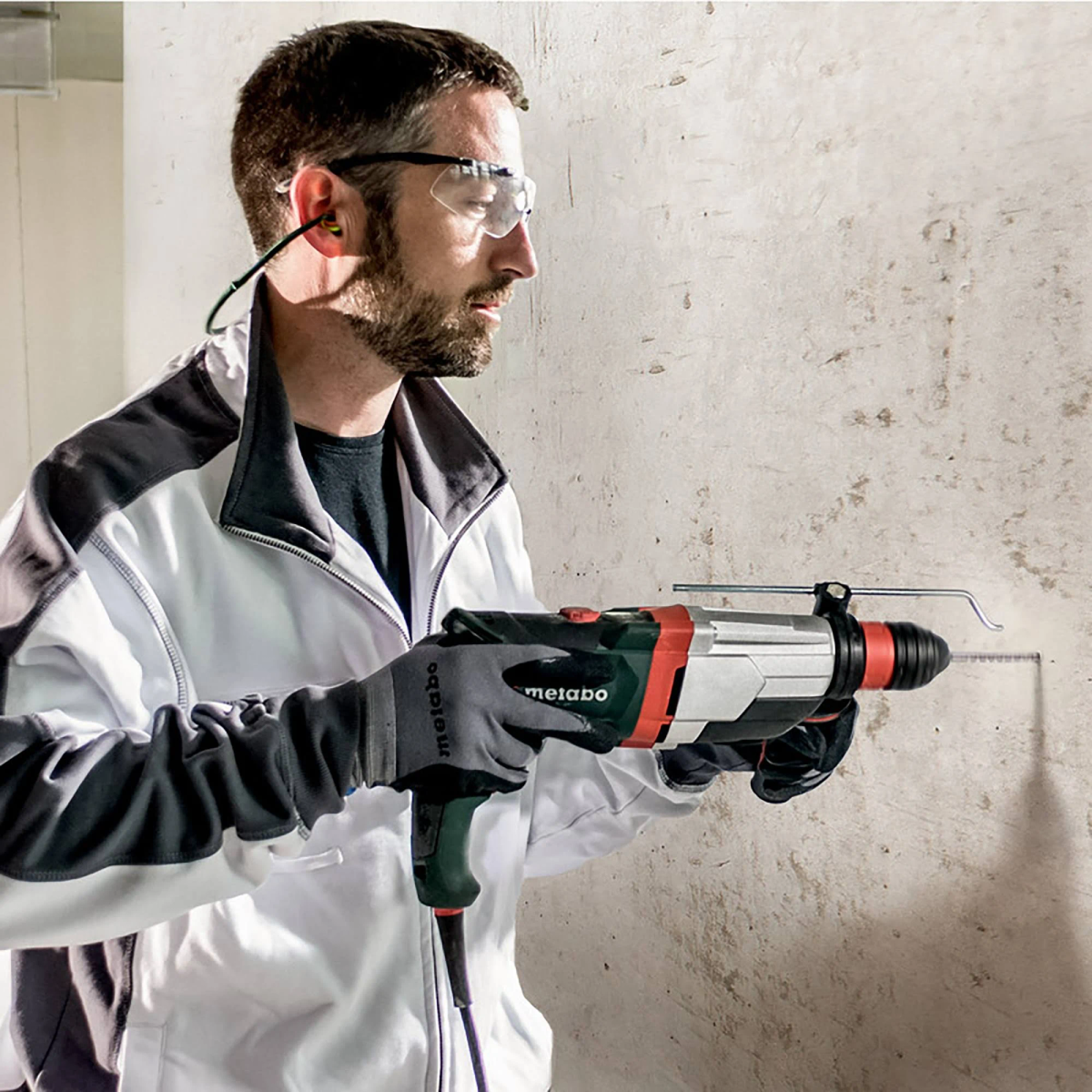Mężczyzna w okularach ochronnych i ochronnikach słuchu wiercący w ścianie.