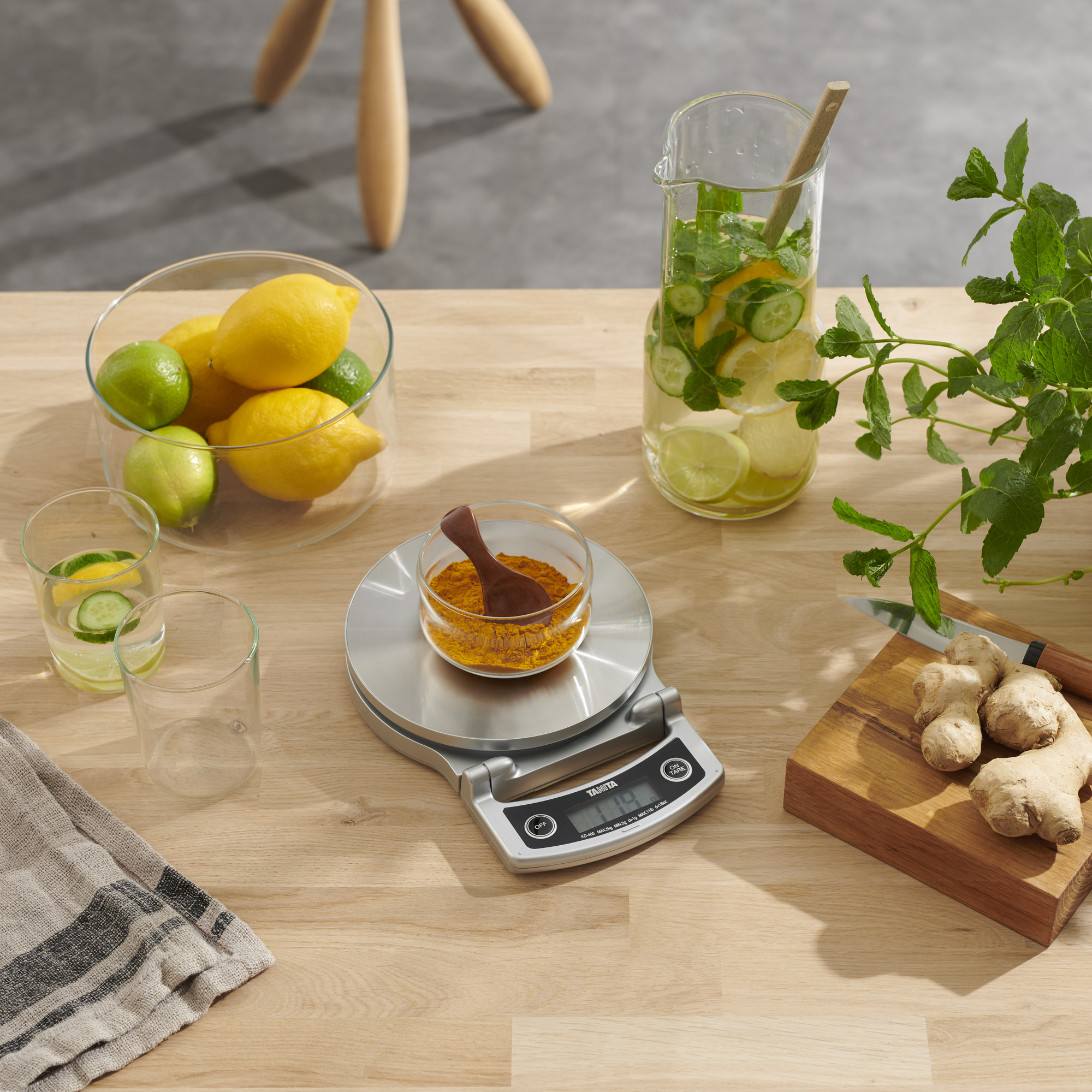 A table with lemons, limes, a scale, a water jar, and herbs. Ginger and a wooden spoon are present.