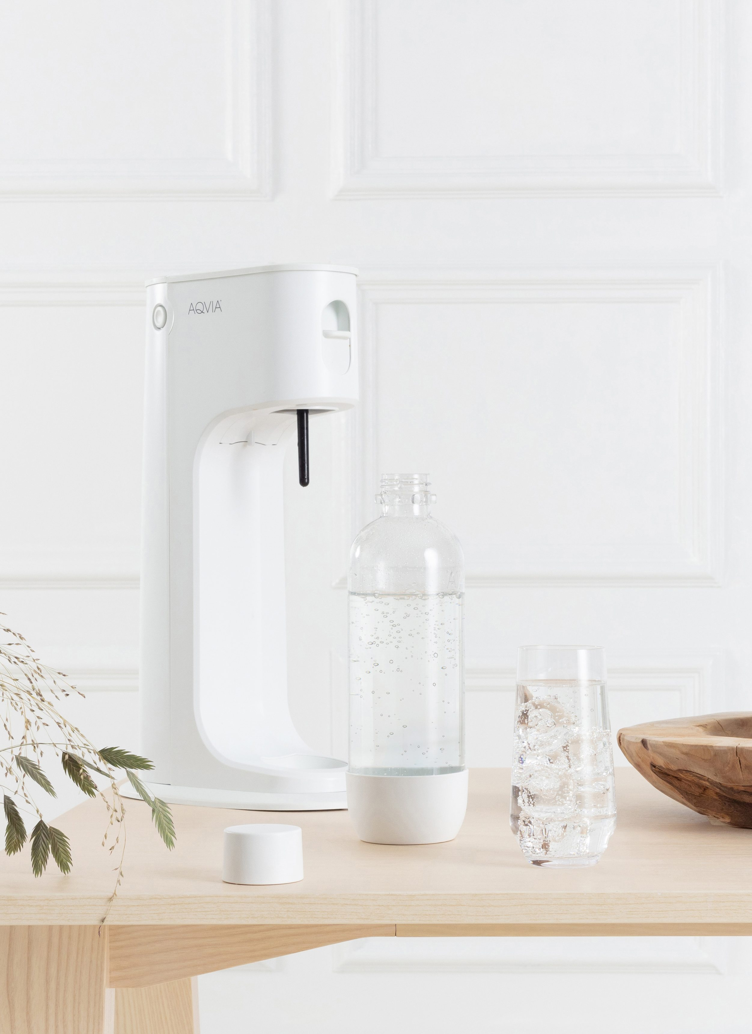 Ein weißer Wasserspender, eine Flasche und ein Glas mit Eis auf einem Holztisch.