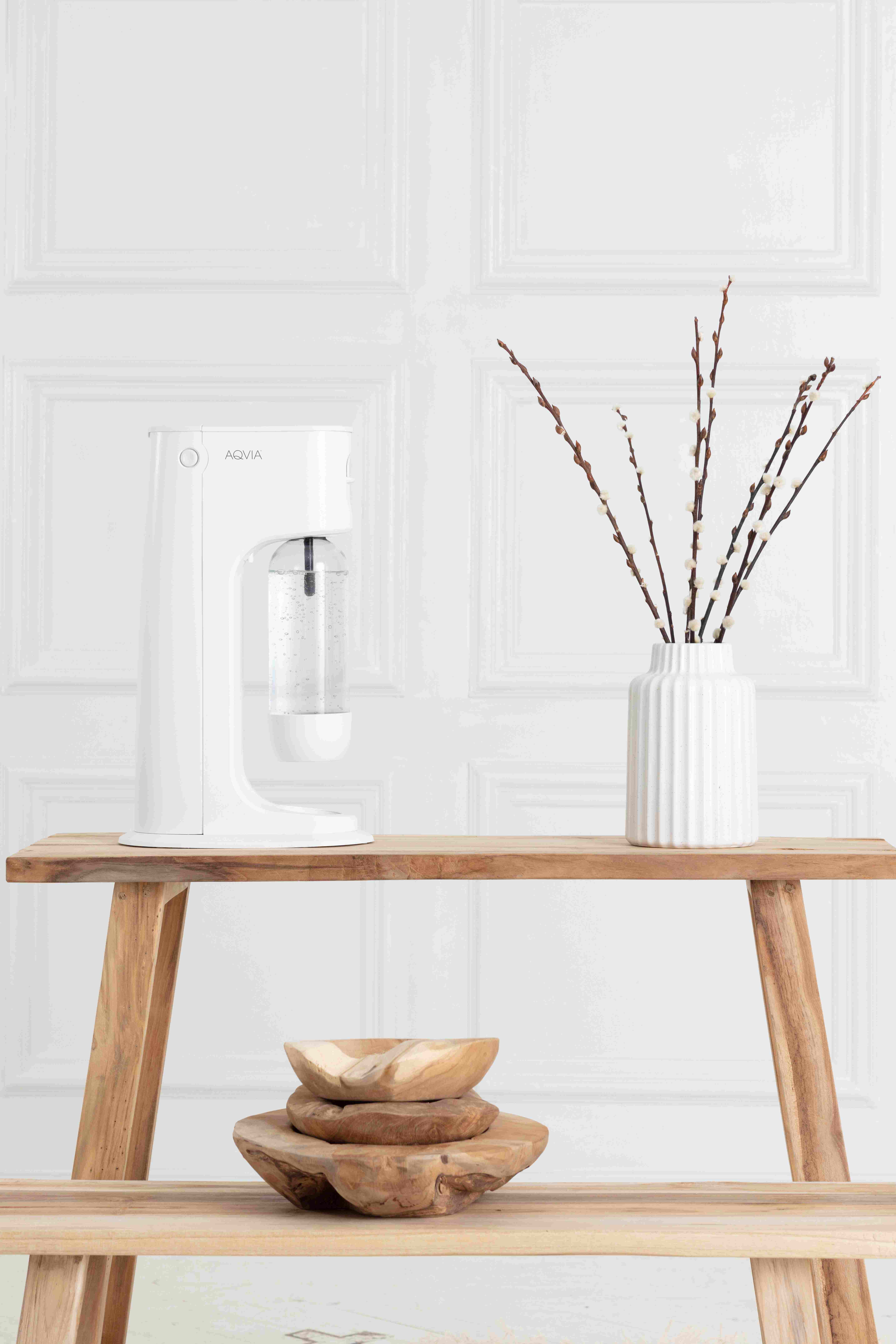 Weißer Sodabereiter auf einem Holztisch mit einer Vase mit Zweigen. Der Hintergrund ist eine weiße Wand.