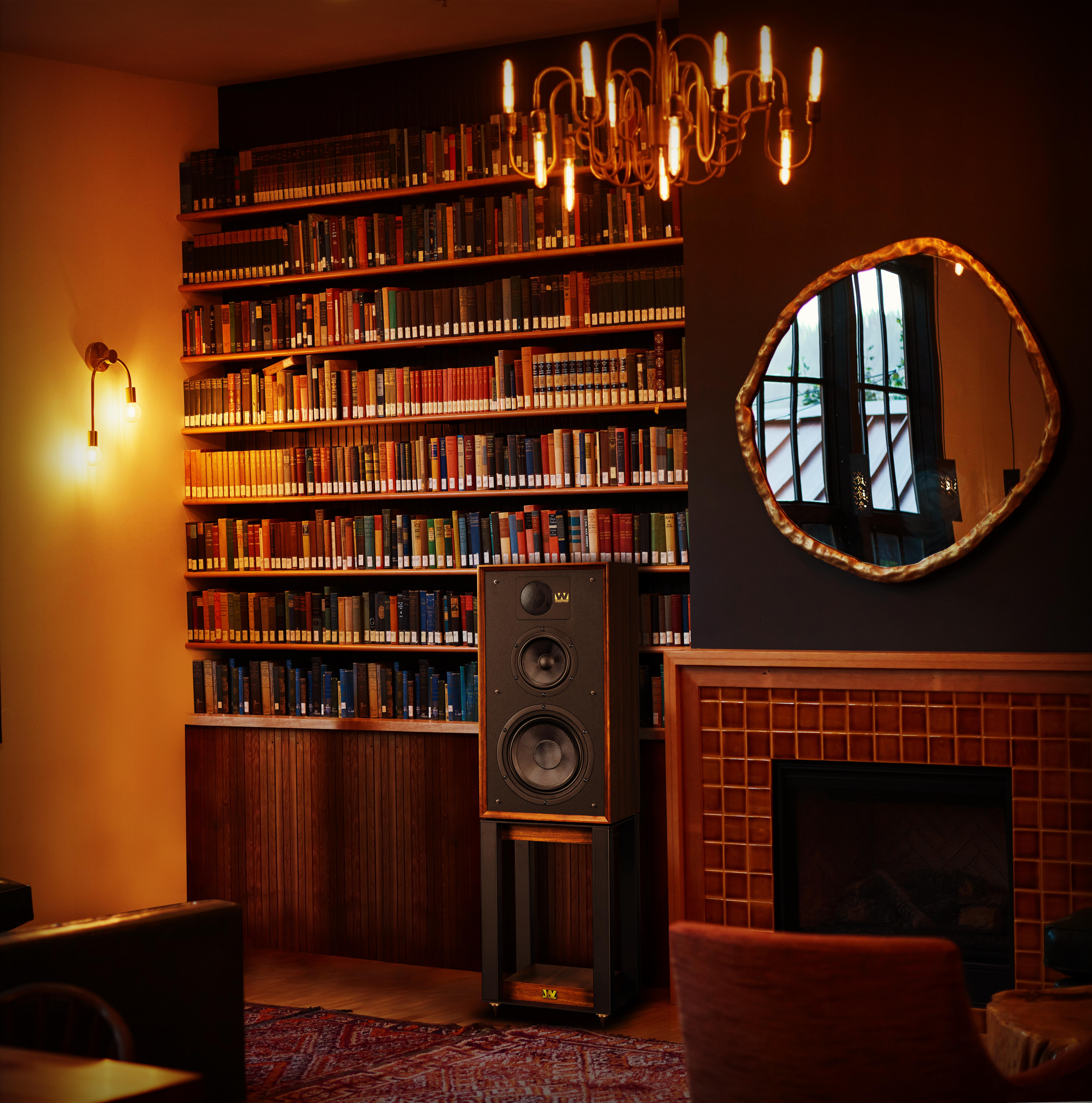 A library interior with a speaker on a stand, fireplace, mirror, chandelier, and bookshelves filled with books.