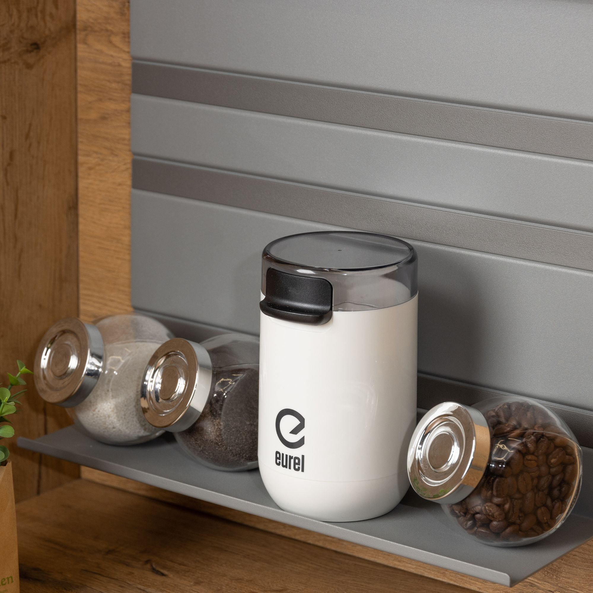 Weiße Kaffeemühle mit transparentem Deckel, neben Gläsern mit Zutaten.