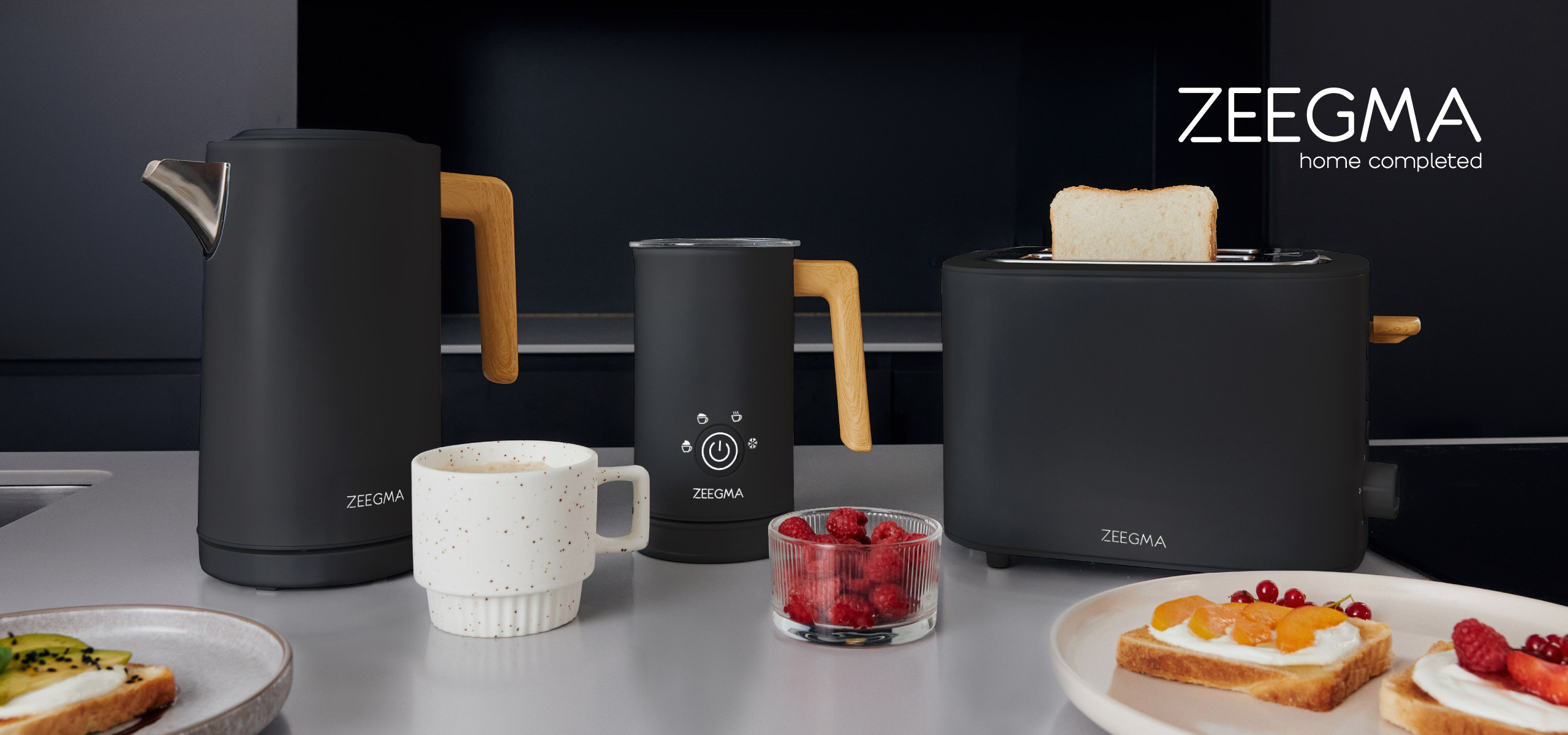 Keuken met zwarte apparaten: waterkoker, melkopschuimer, broodrooster en een mok. Eten en logo zichtbaar.