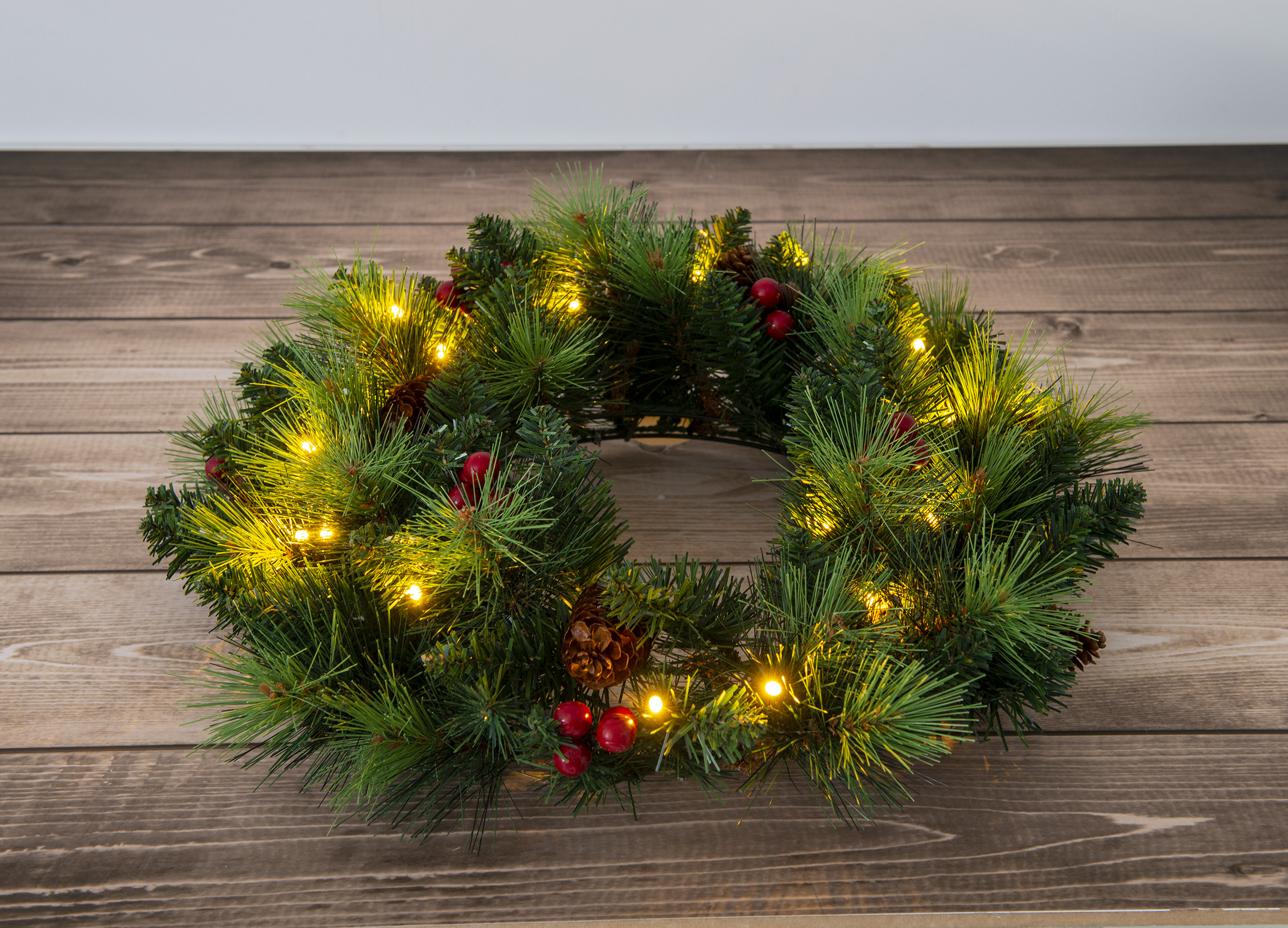 Weihnachtskranz mit Lichtern, Tannennadeln, roten Beeren und Zapfen, auf Holz.