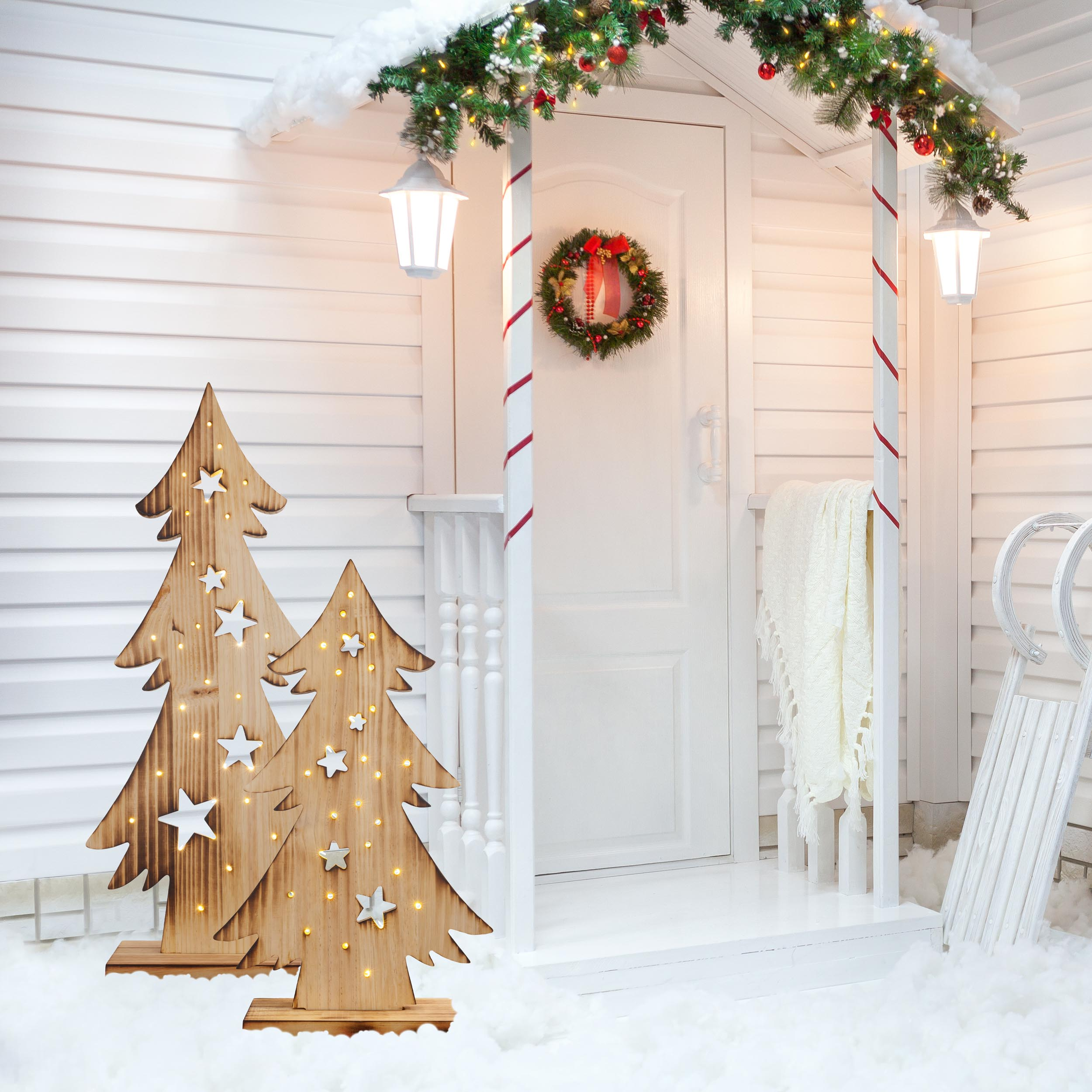 Weihnachtsbaumdekorationen aus Holz mit Lichtern auf einer Veranda mit weißer Tür und Schnee.