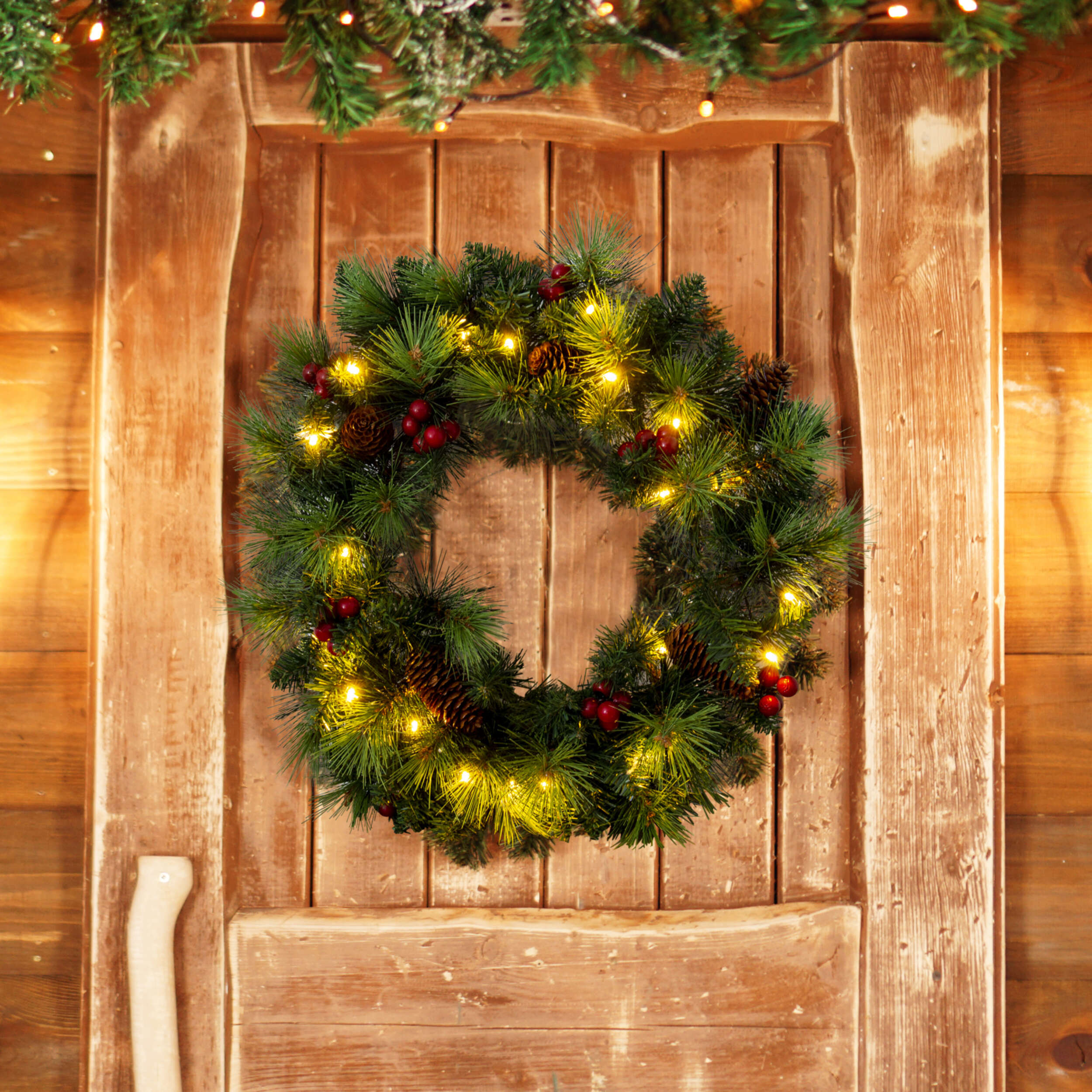Ein Weihnachtskranz mit Lichtern an einer Holztür. Tannenzapfen und rote Beeren sind sichtbar.
