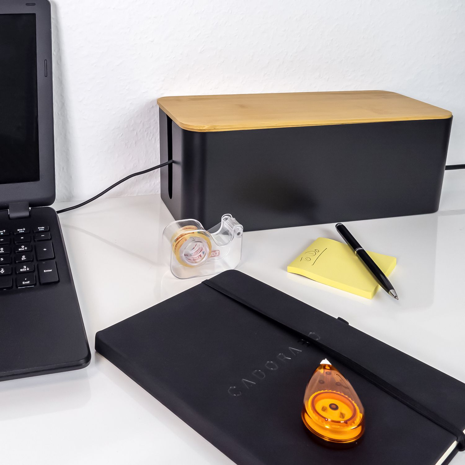 Schreibtisch-Setup: Laptop, Kabelbox, Klebebandspender, Notizblock, Stift und Notizbuch.