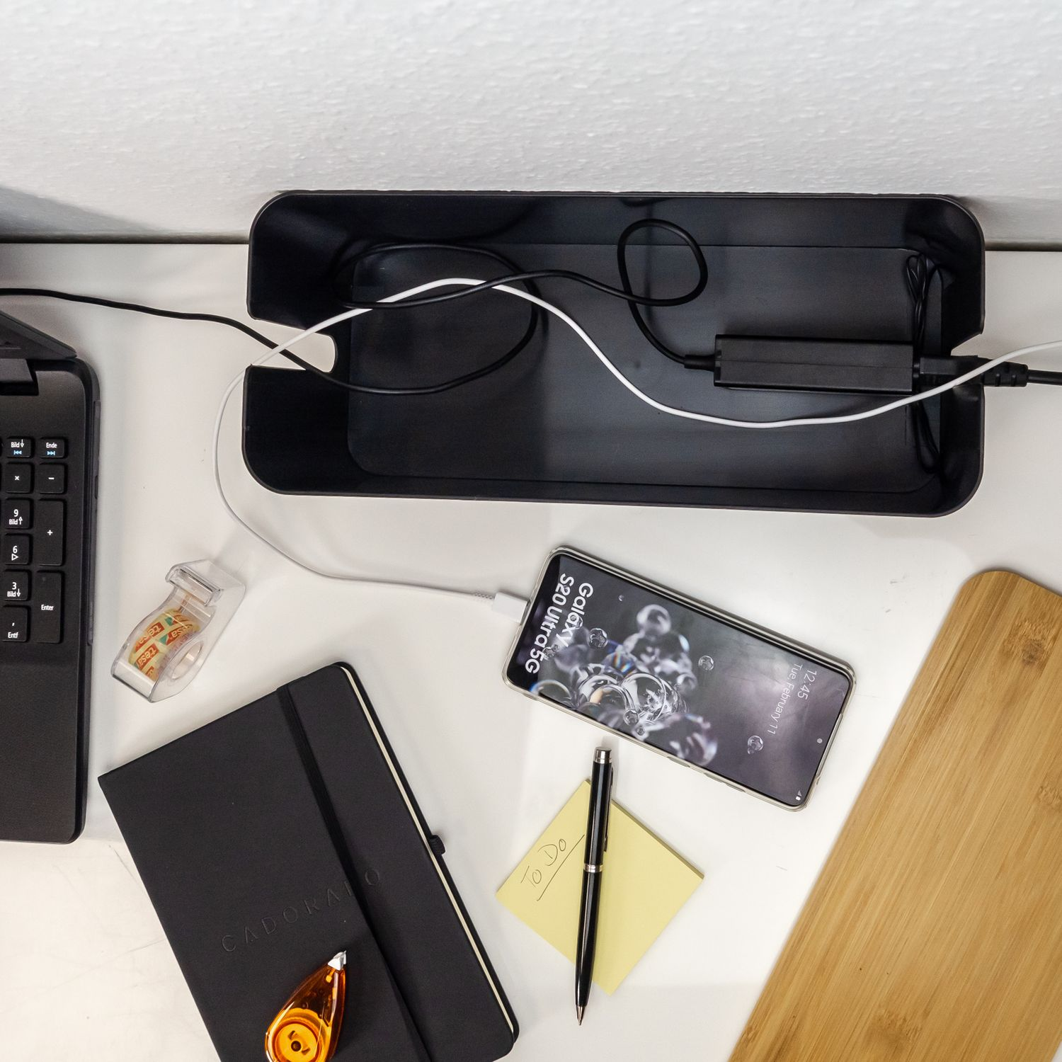 Schreibtischszene mit einem Telefon, das in einer schwarzen Kabelbox, einem Notizblock, einem Stift, einem Klebebandspender und einem Schneidebrett geladen wird.