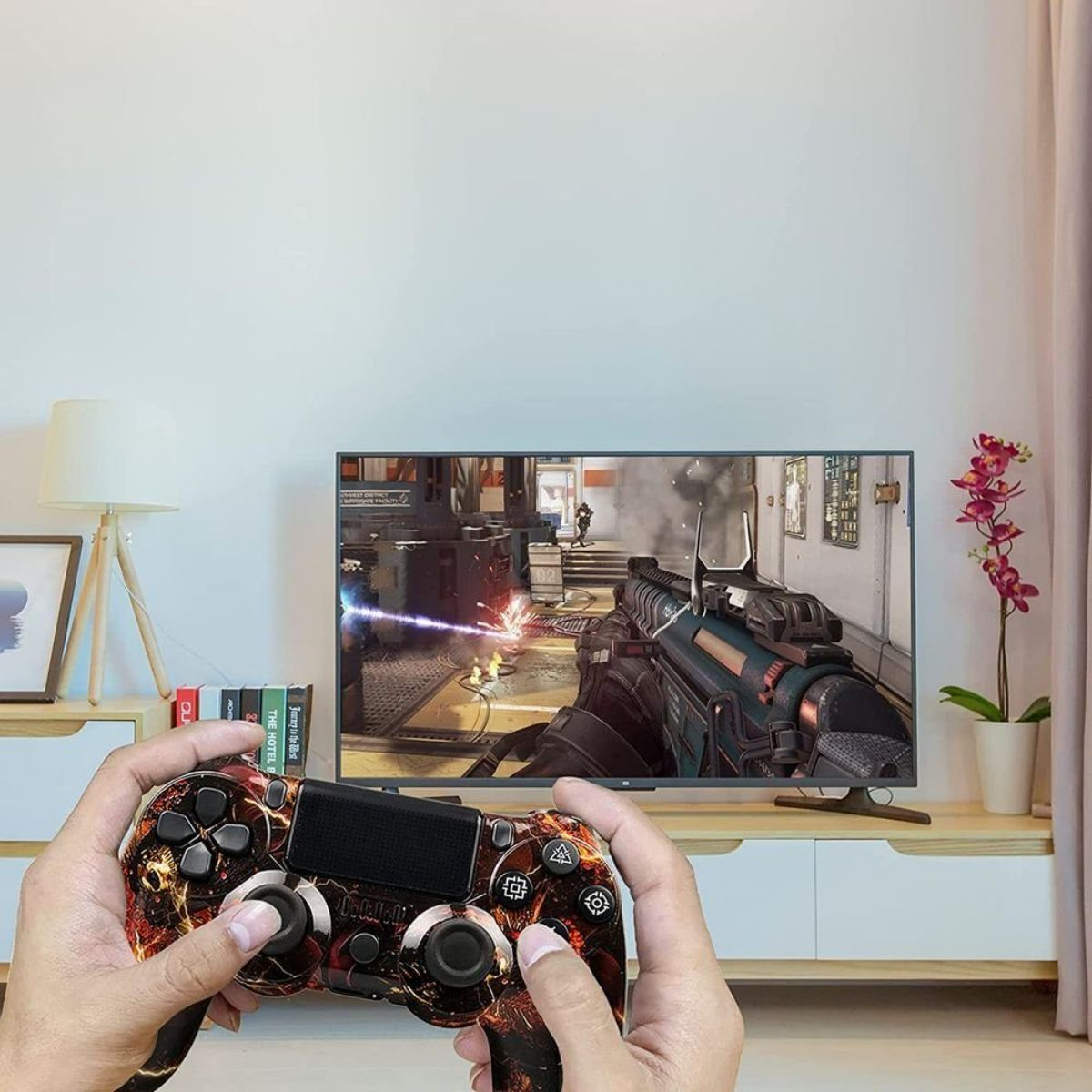 Manos sosteniendo un mando de juego con diseño, jugando en un televisor. La habitación tiene una lámpara y una ventana.