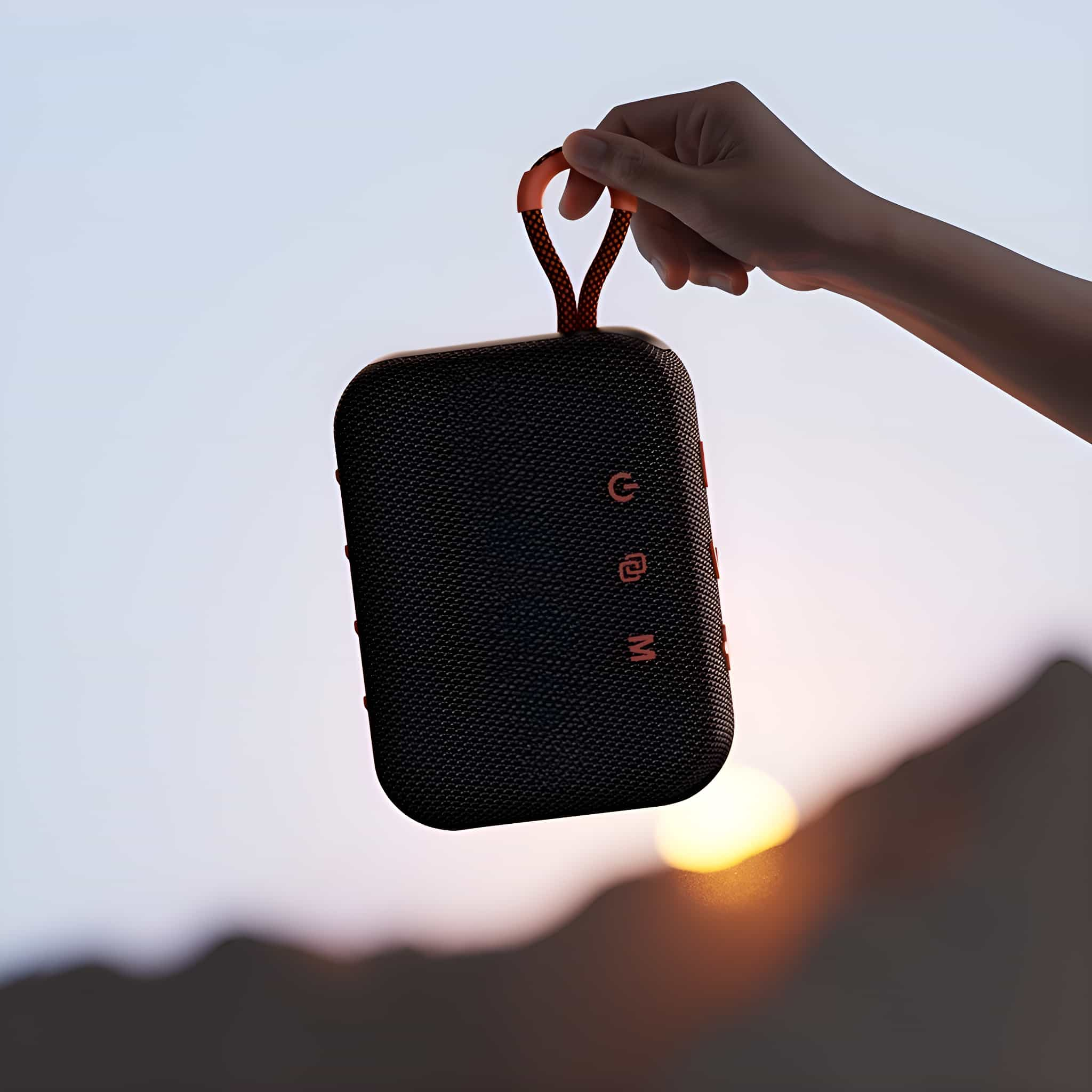 Hand hält einen schwarz-orangenen Lautsprecher mit rotem Riemen vor Sonnenuntergang.