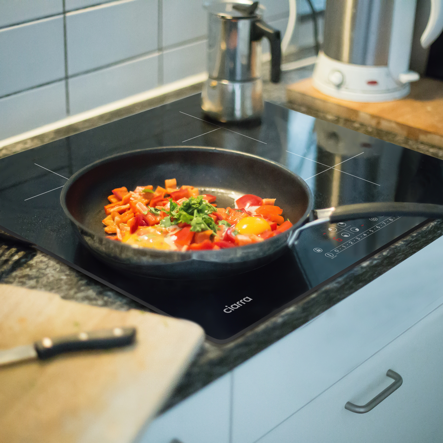 Eine Bratpfanne mit Eiern und Gemüse steht auf einem schwarzen Kochfeld. Küchenzubehör im Hintergrund.