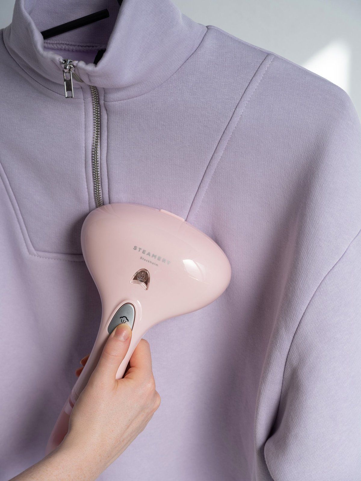 Eine Hand, die einen lavendelfarbenen Sweatshirt mit einem rosa Dampfgarer dämpft.