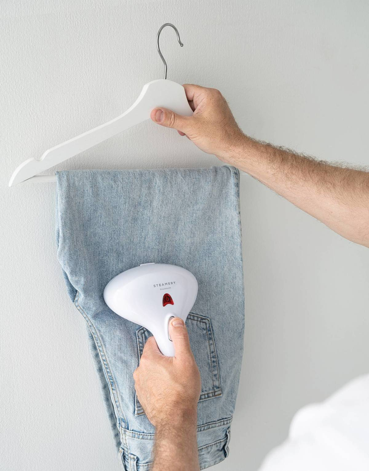Person bügelt Jeans mit einem weißen Steamer an einem weißen Kleiderbügel vor einer weißen Wand.
