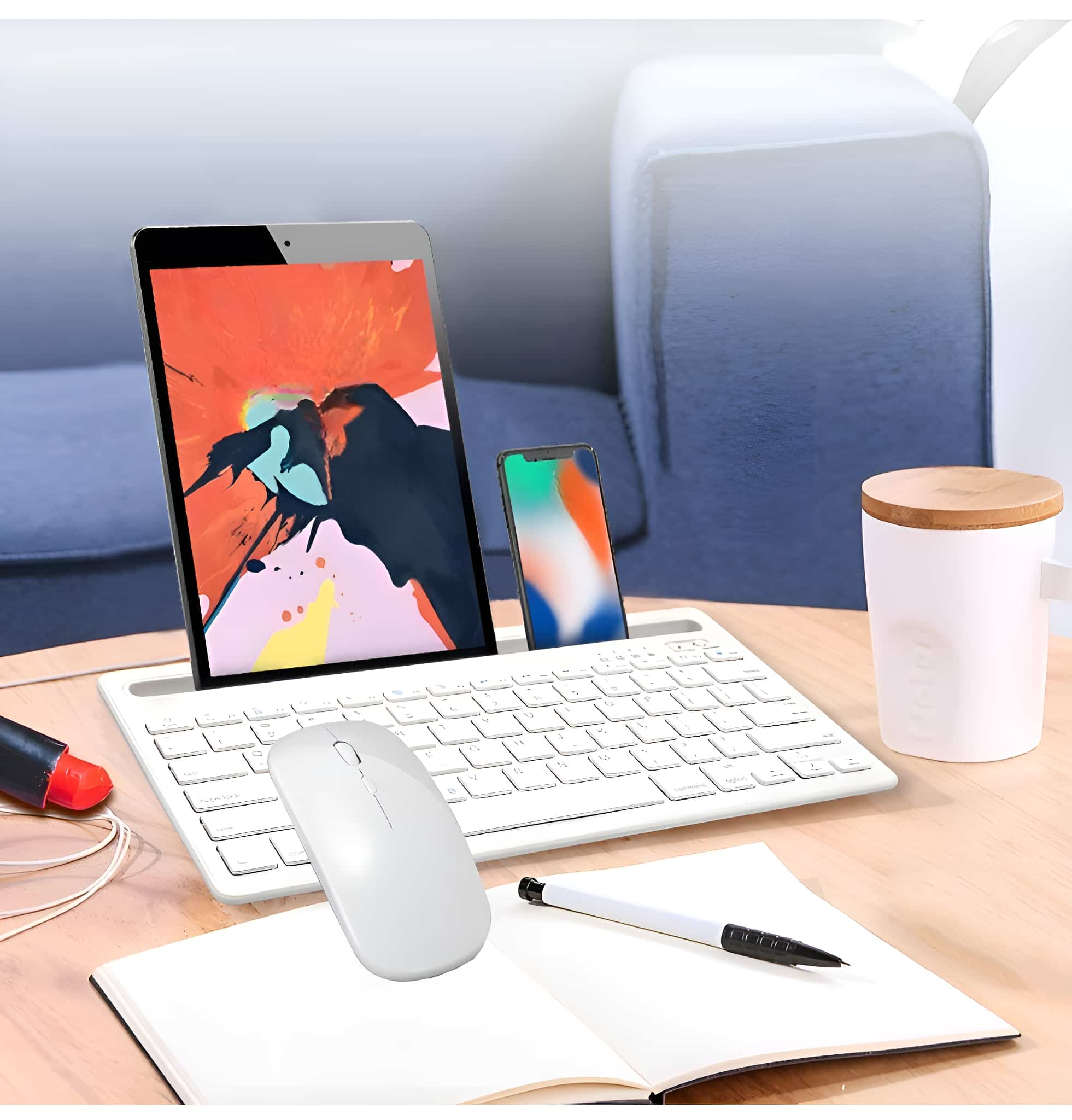 Tablet, Telefon, Tastatur, Maus und Notizblock stehen auf einem Schreibtisch mit einem Sofa im Hintergrund.