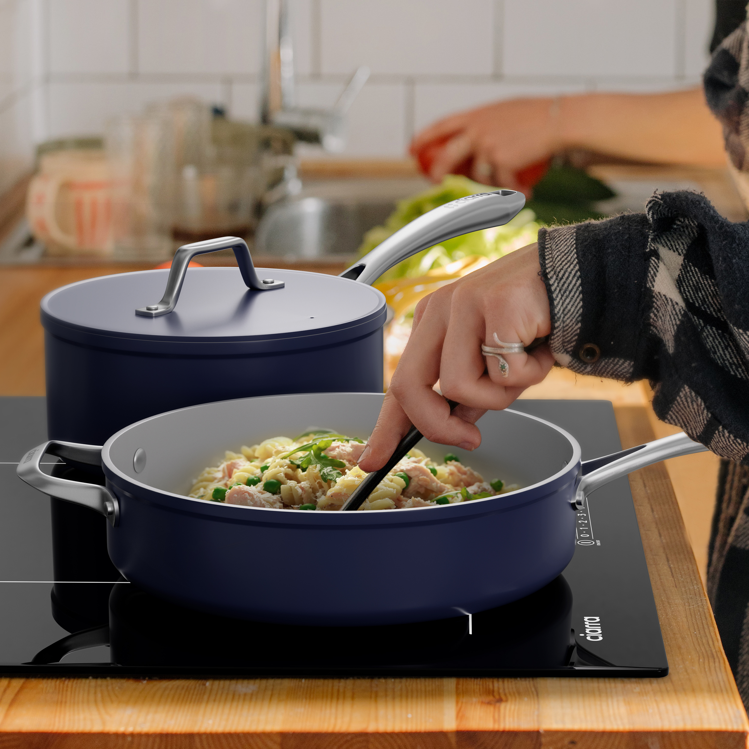 A person cooking pasta in a blue pan on a black stovetop. A pot and background elements.