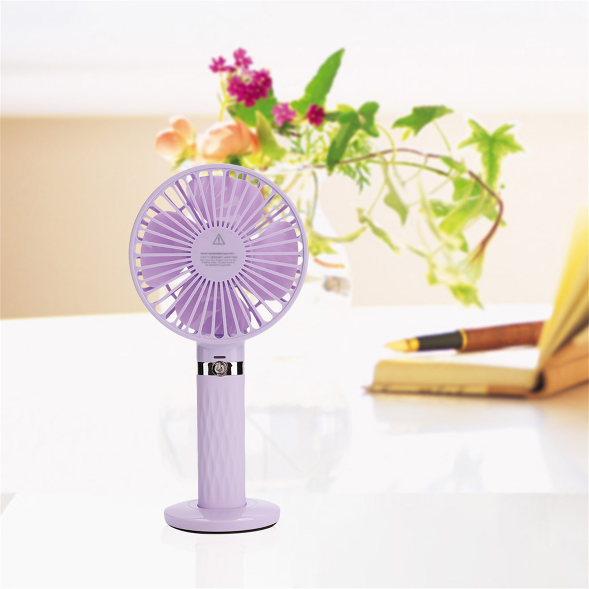 Lila Handventilator auf einem weißen Tisch. Ein Buch und Blumen sind im Hintergrund.