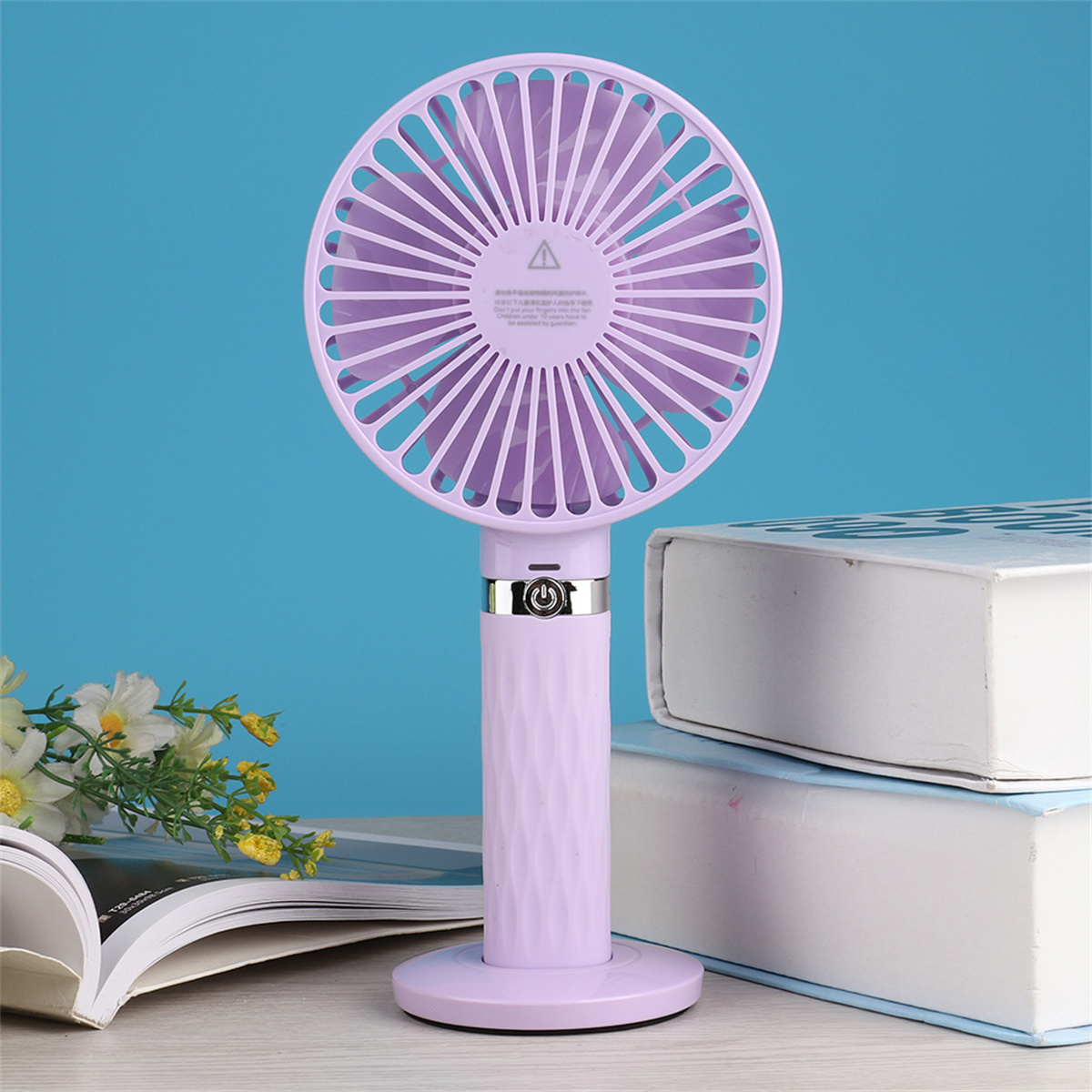 Ein violetter tragbarer Ventilator steht auf einem Tisch neben Büchern. Der Ventilator hat einen weißen Sockel und Griff.