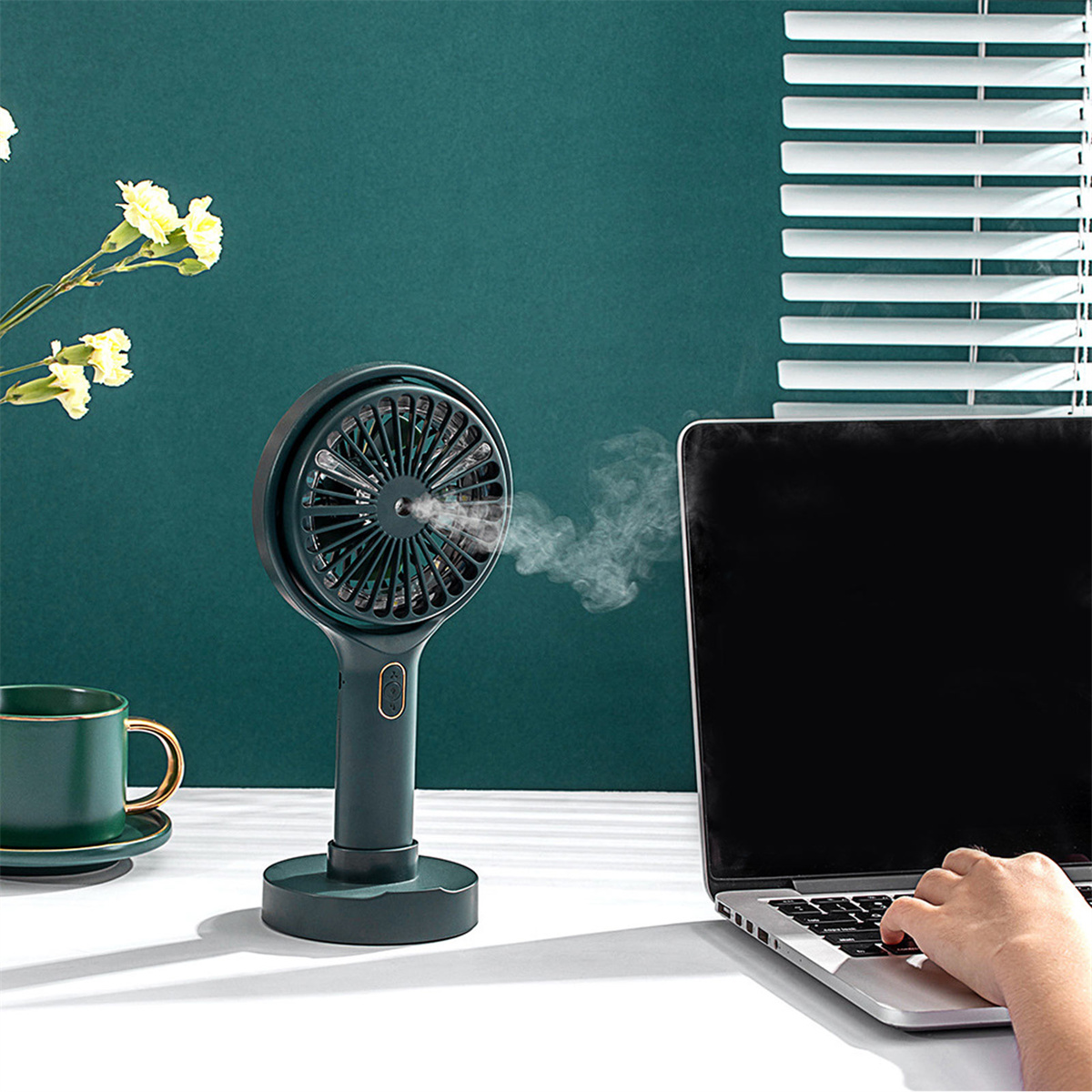 Ein dunkelgrüner tragbarer Ventilator steht auf einem weißen Tisch und bläst Luft in Richtung eines Laptops.