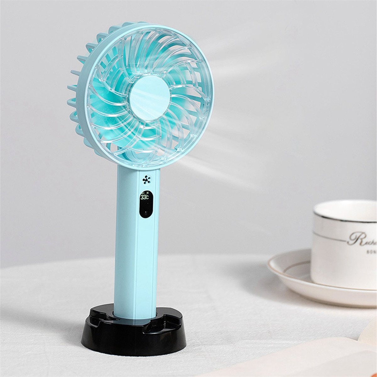 Blauer Handventilator mit schwarzem Sockel, auf einem Tisch mit Tasse und Teller.