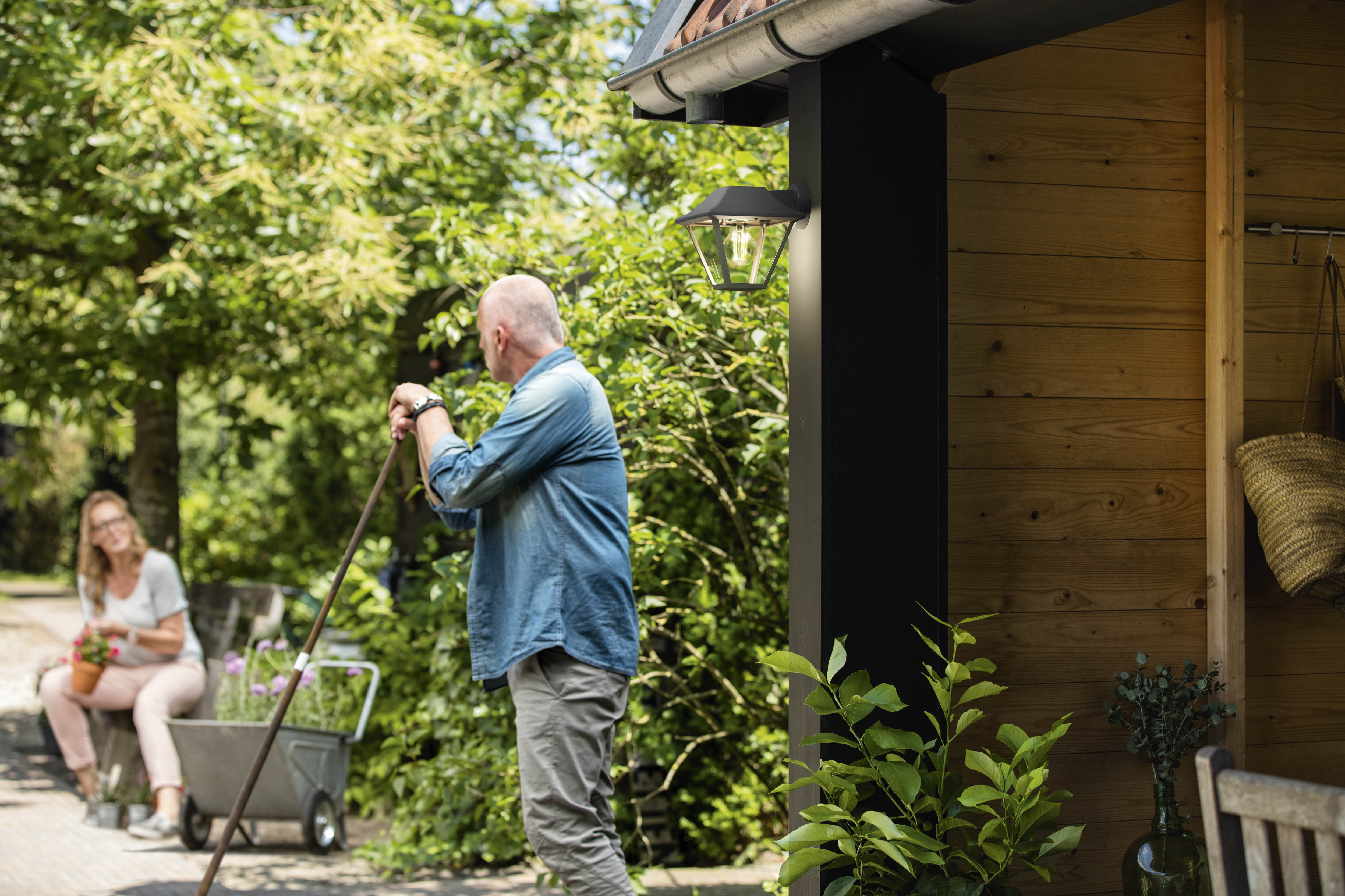 Ein Mann mit einer Harke und eine Frau mit einem Topf in einer Gartenszene mit einem Holzschuppen.